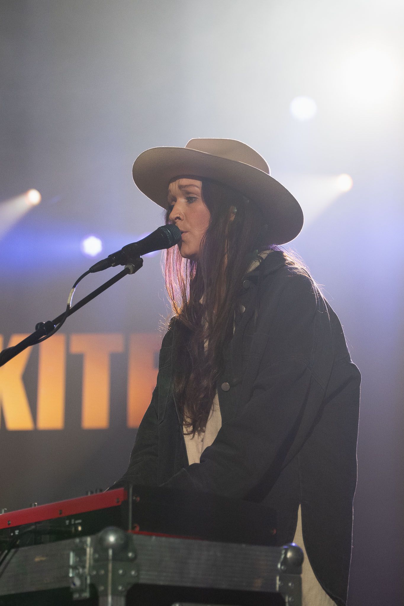 Performer in wide-brimmed hat singing at keyboard during concert performance.