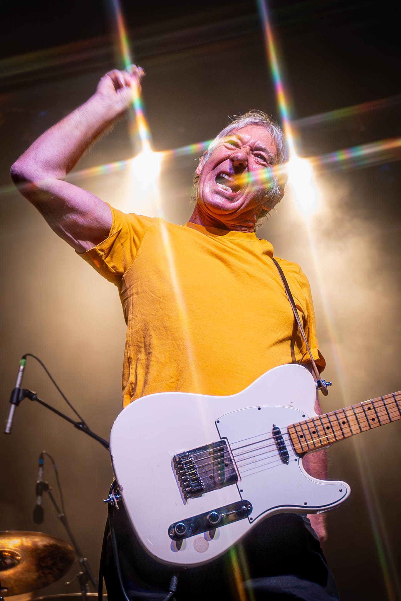 Musician from The Buzzcocks in yellow shirt plays white electric bass guitar under bright stage lights.