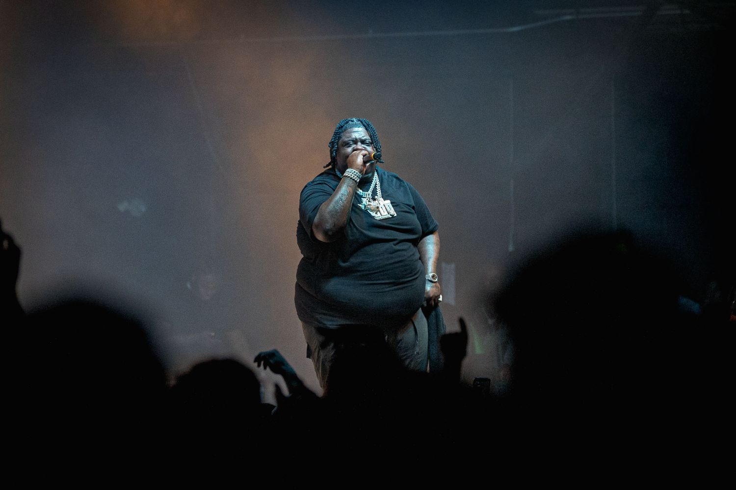 Dramatic spotlight illuminates BigXthaPlug on darkened stage with crowd silhouettes visible.