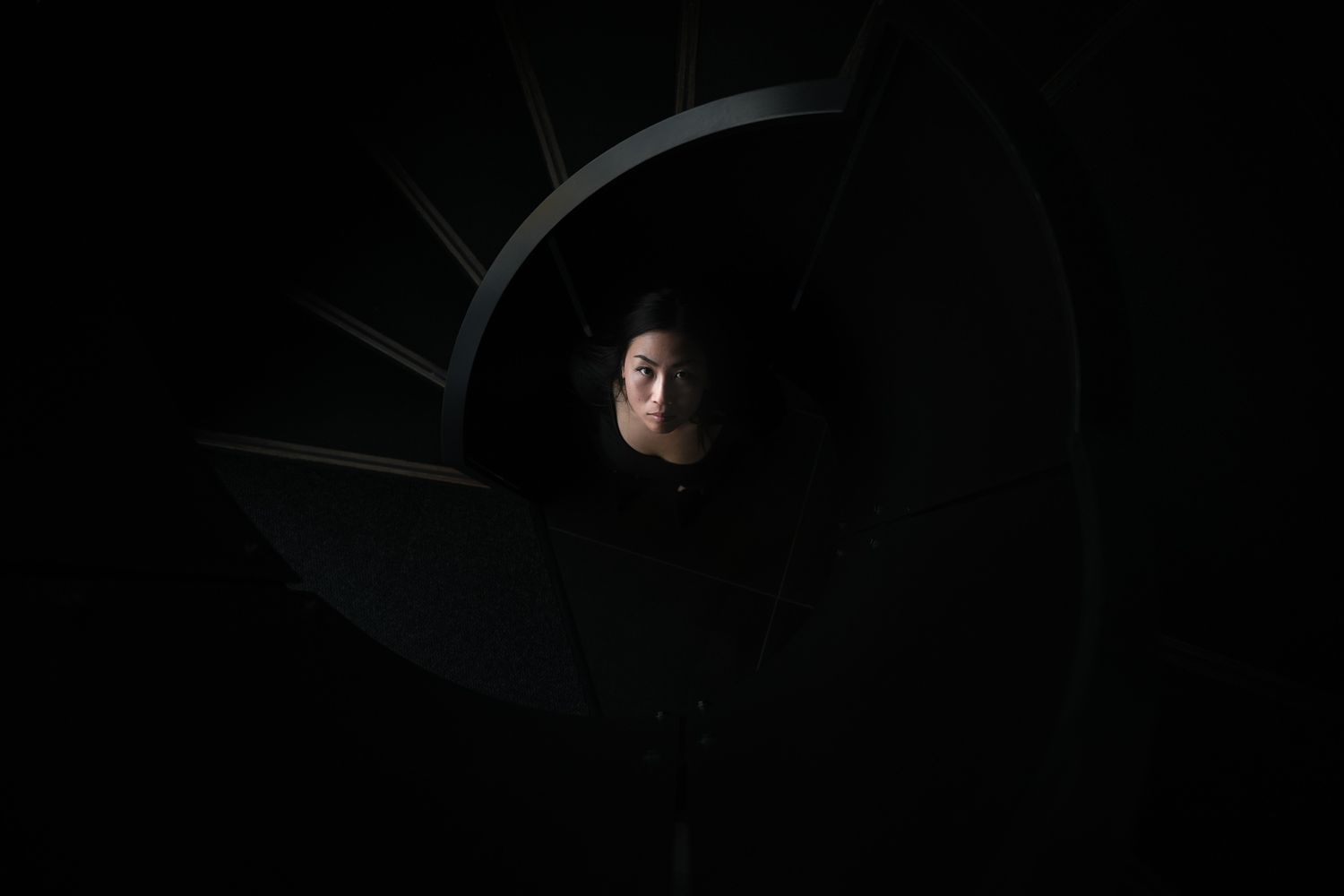 A person's face emerges from darkness through a circular opening of a spiral staircase with geometric lines creating a dramatic contrast.