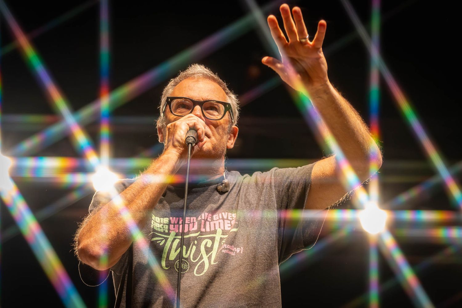 Stage performer raises arms during concert with dramatic prismatic lighting effects.