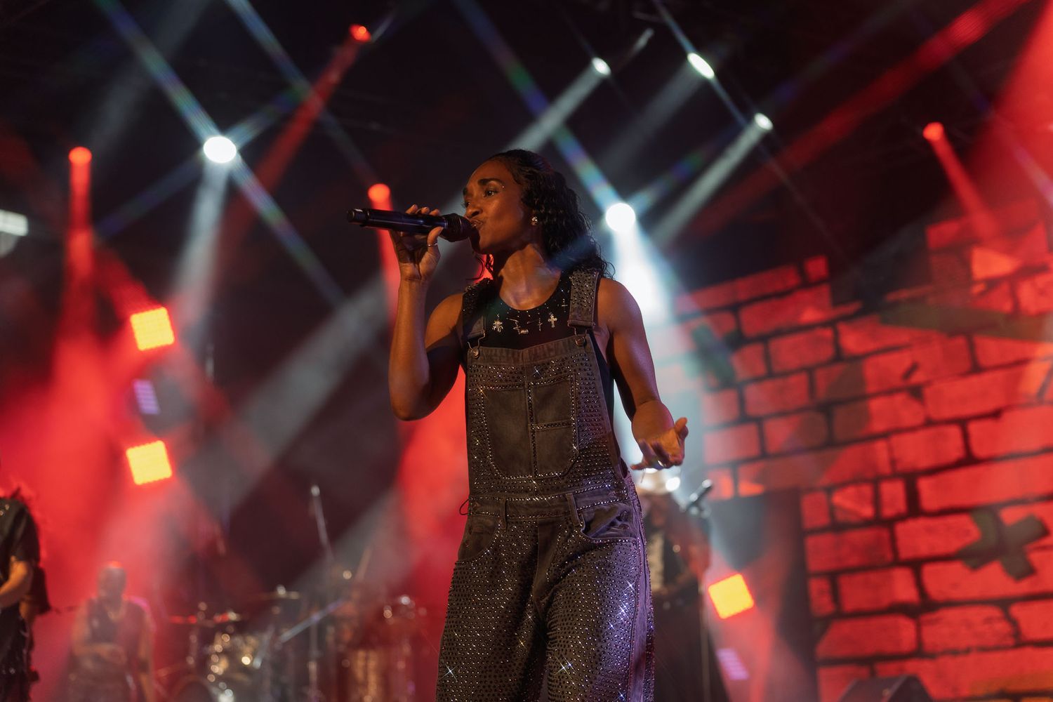 Performer in overalls singing on stage with dramatic red lighting and brick backdrop.