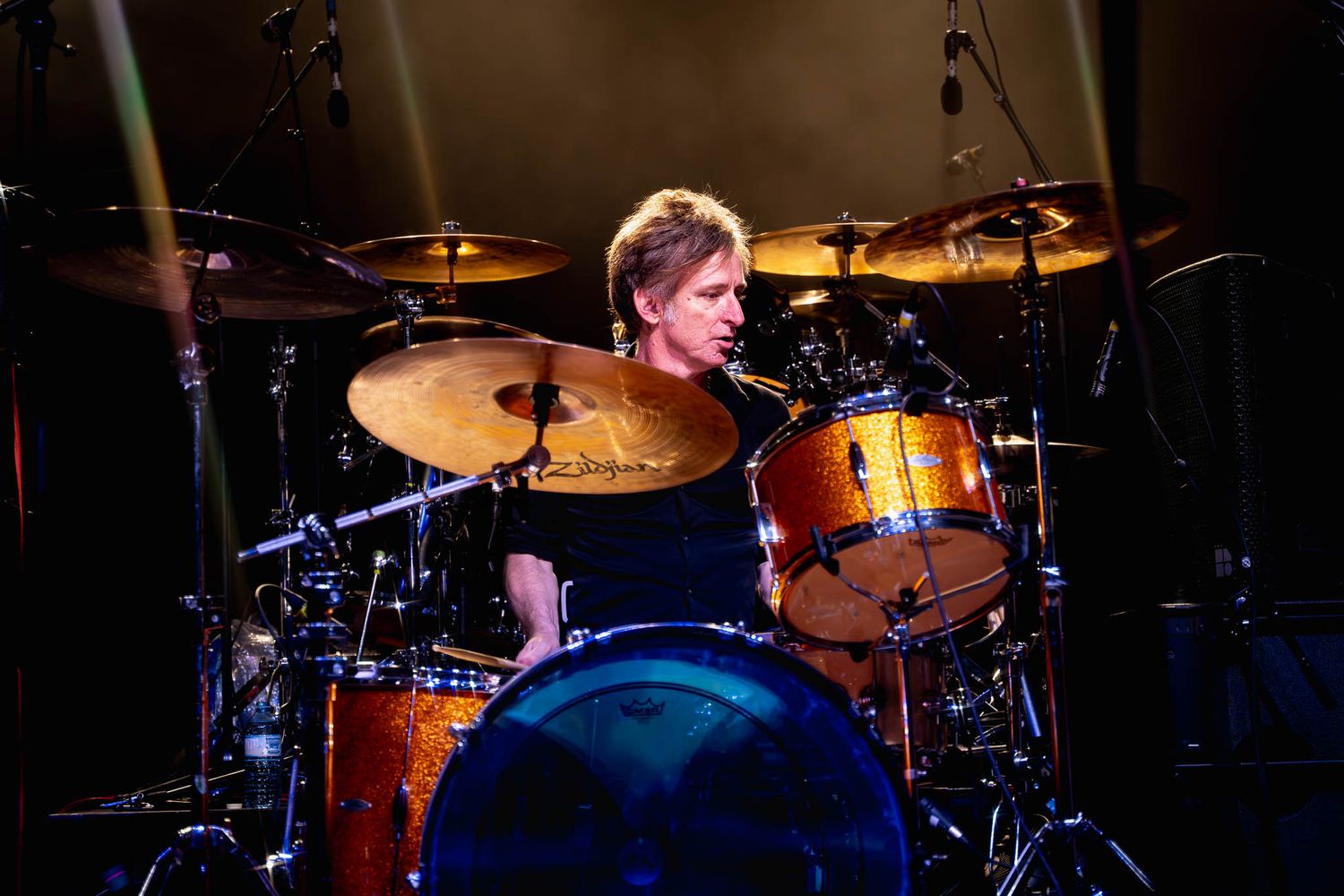 Drummer performing behind drum kit on dimly lit concert stage.