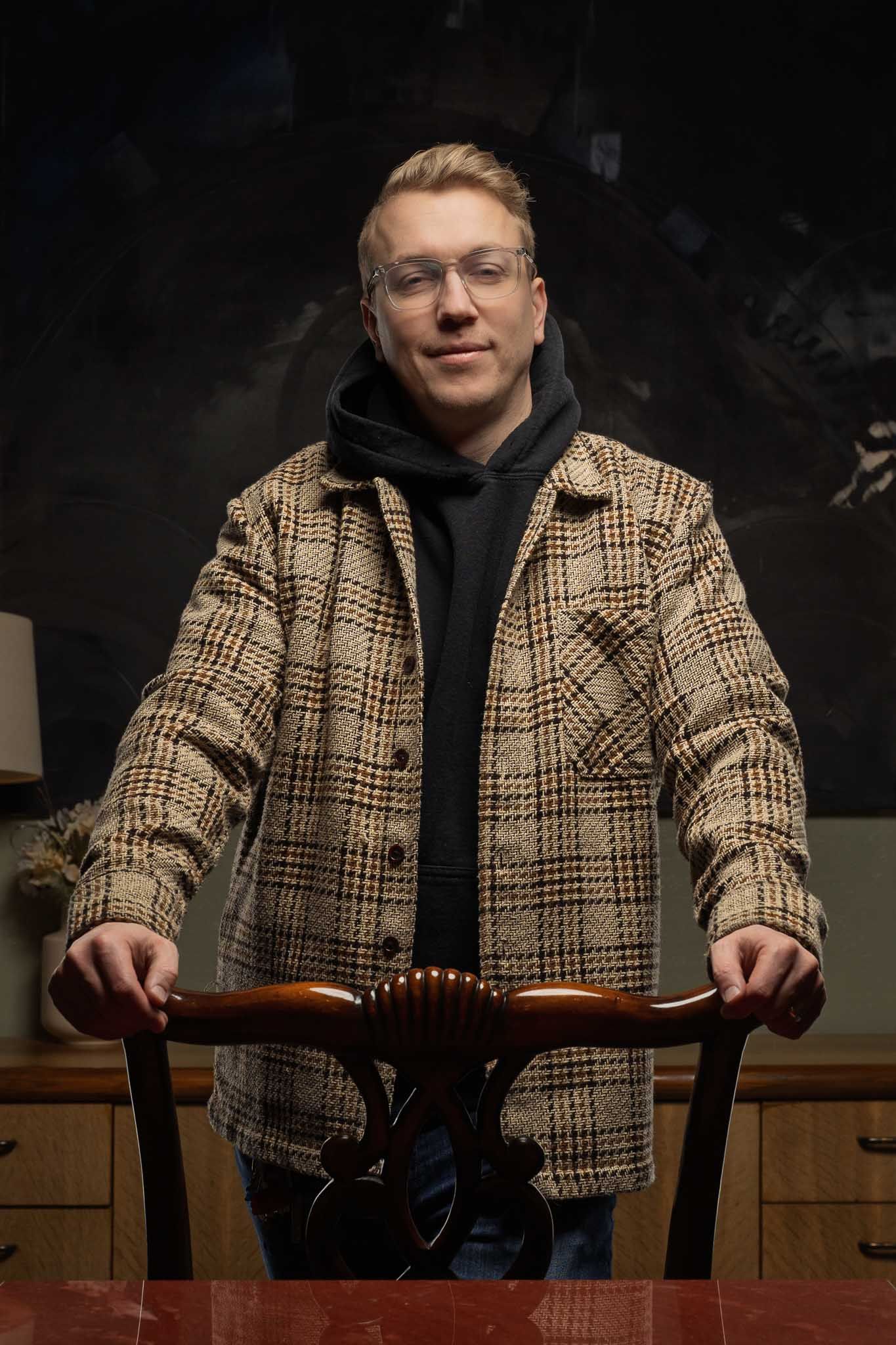 Person wearing checkered coat and glasses sits casually at wooden table.