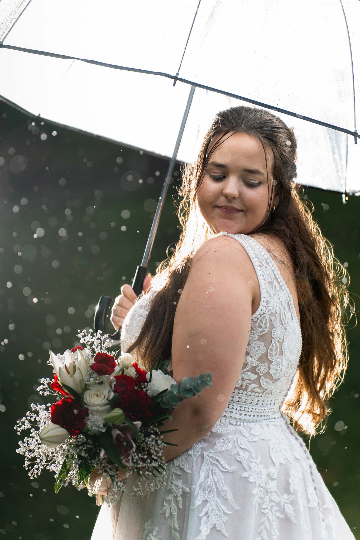 Bride with clear umbrella and burgundy bouquet poses in rain.