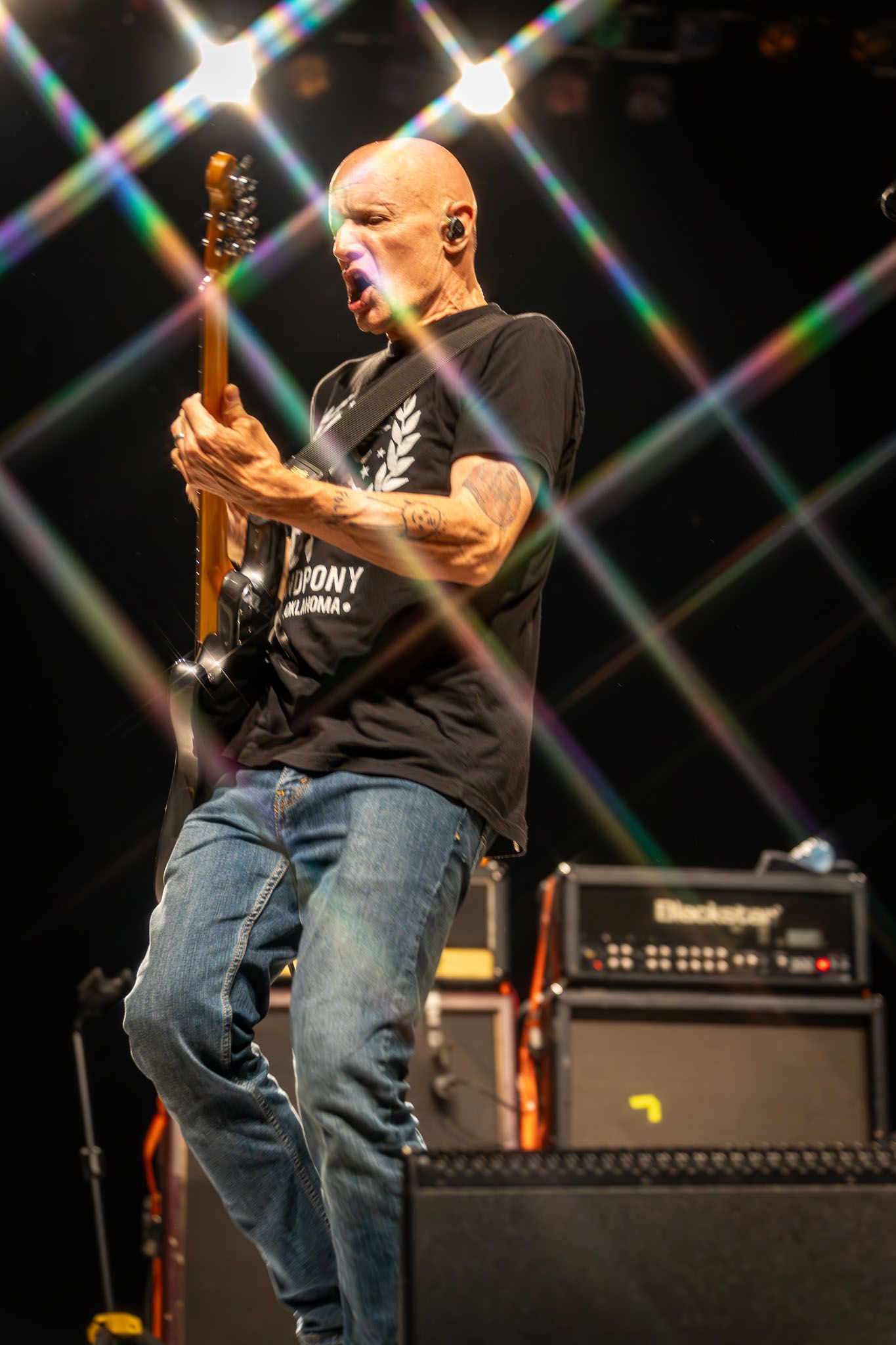 Guitarist in black t-shirt and jeans playing electric guitar on stage with amp setup.