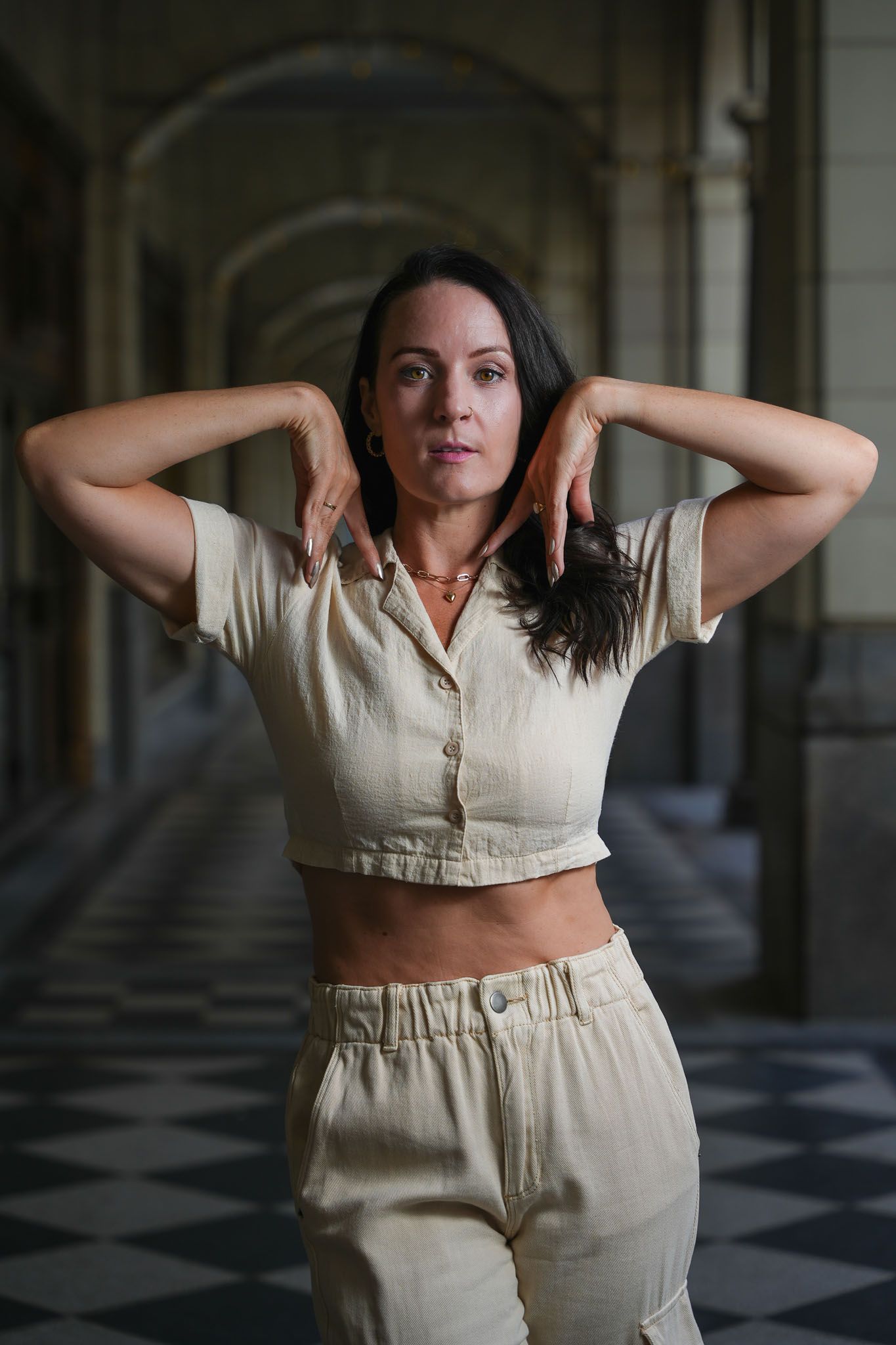 Dancer in cream colored outfit performs expressive arm movements in checkered hallway.