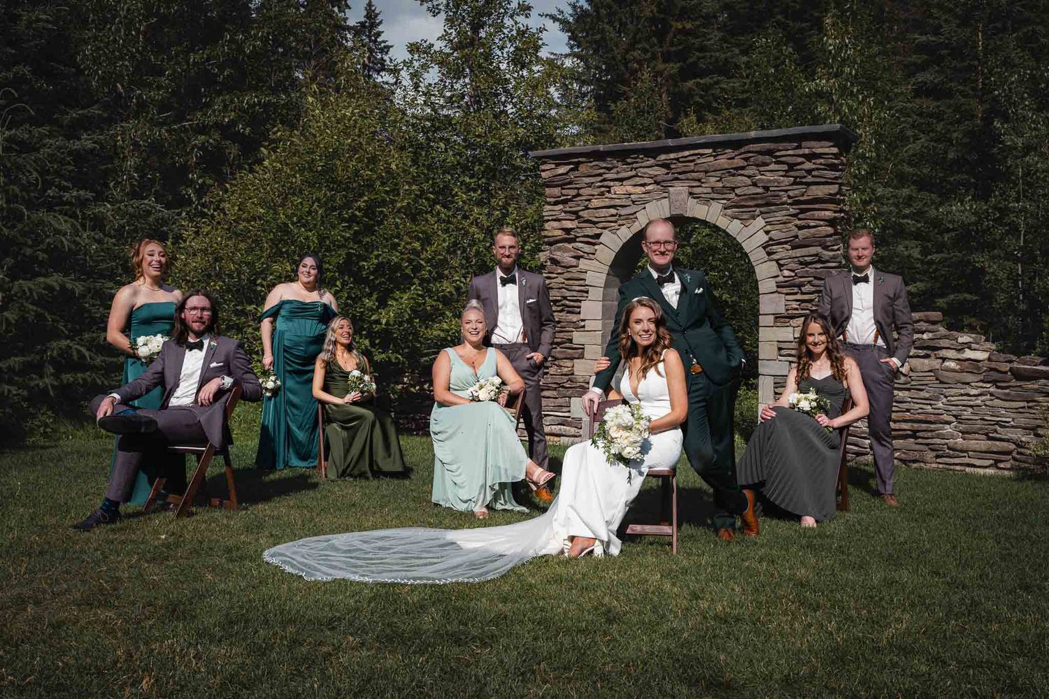 Wedding party relaxes on a stone wall surrounded by lush greenery during an outdoor celebration.
