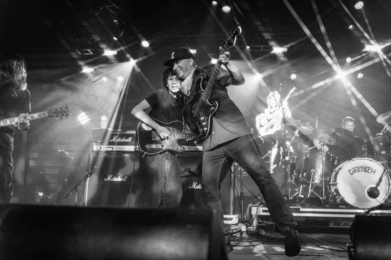 Tom Morello and son Roman Morello performs on dimly lit stage with spotlights creating dramatic silhouettes during live show.