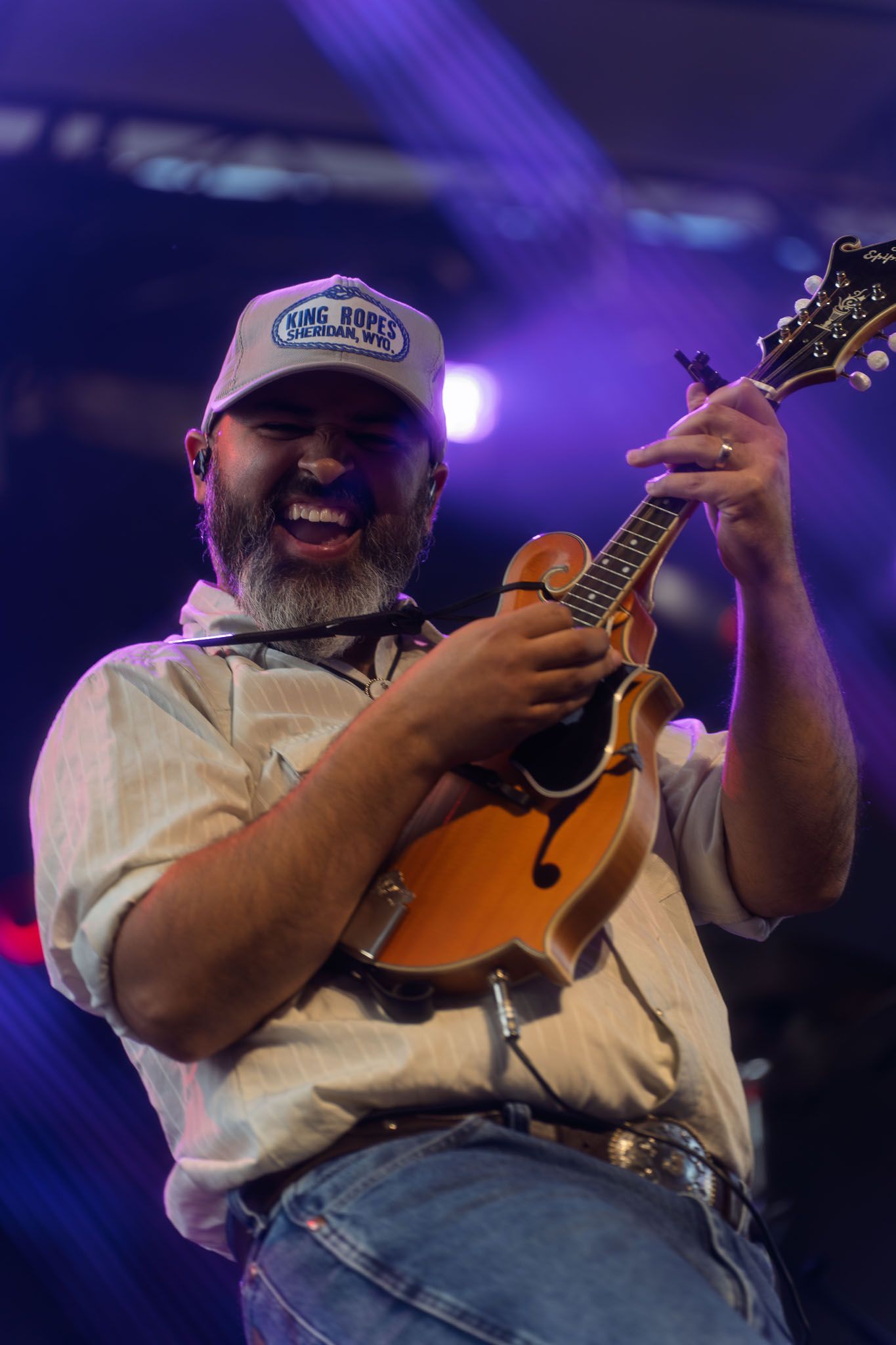 Musician playing mandolin while wearing baseball cap under purple stage lighting.