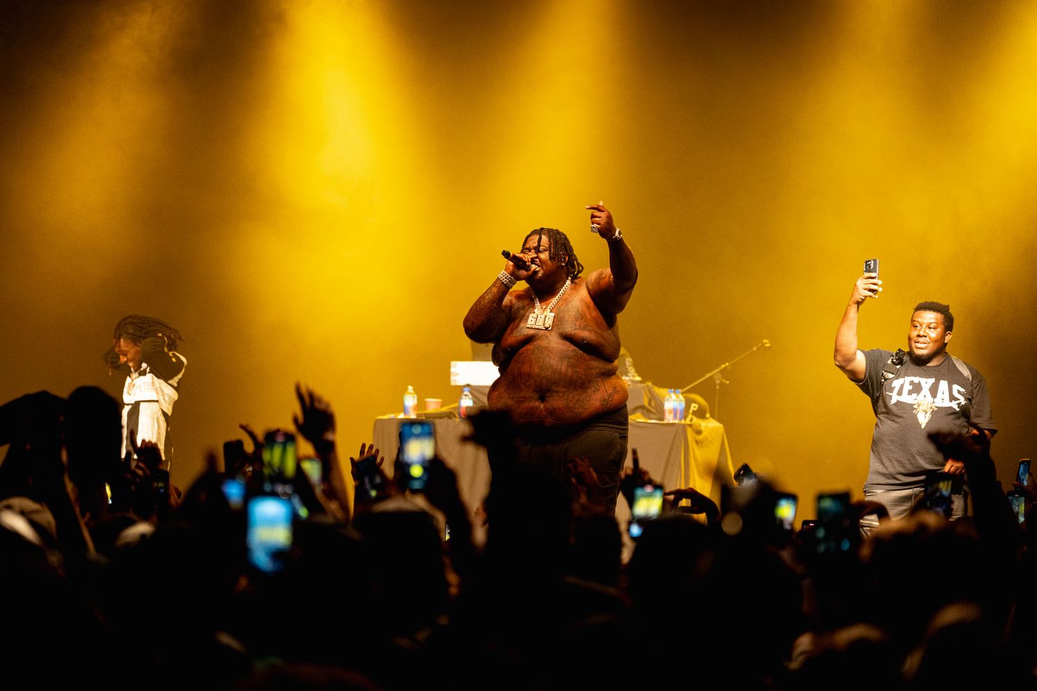 BigXthaPlug on stage with golden lighting and enthusiastic crowd raising phones.