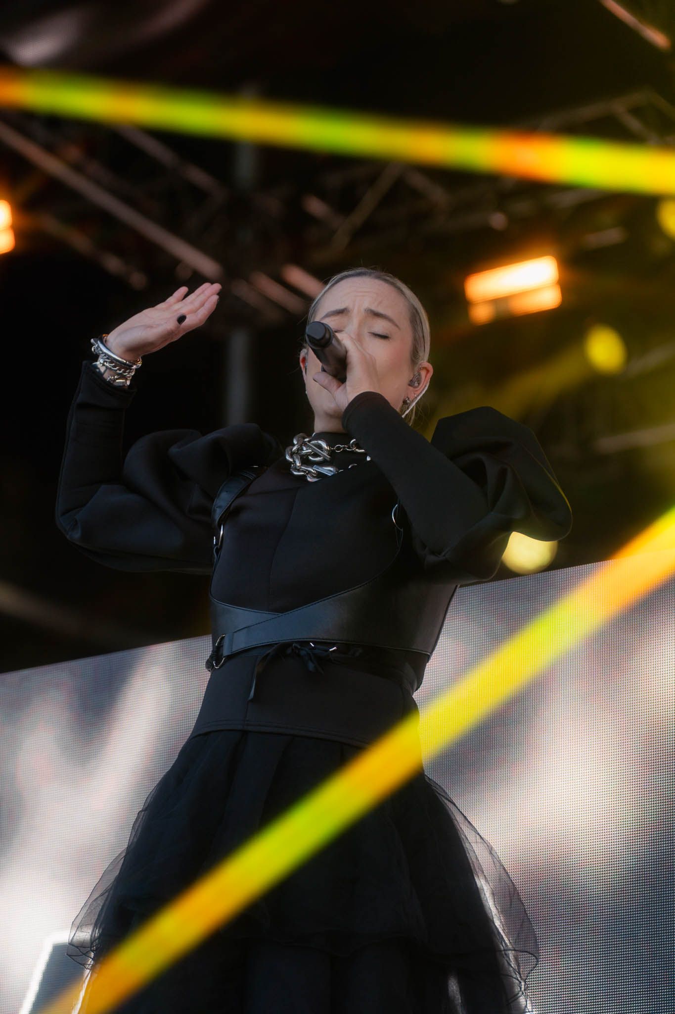 Hayla in black outfit singing on stage with dramatic yellow lighting effects.