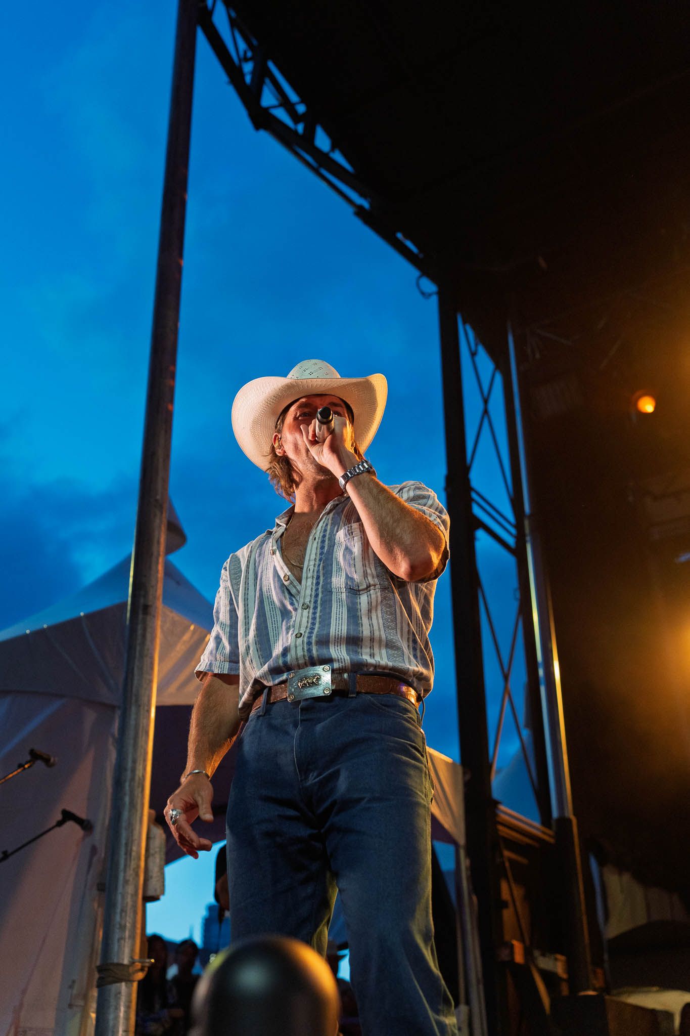 Performer in western wear stands on outdoor stage at dusk with dramatic lighting.