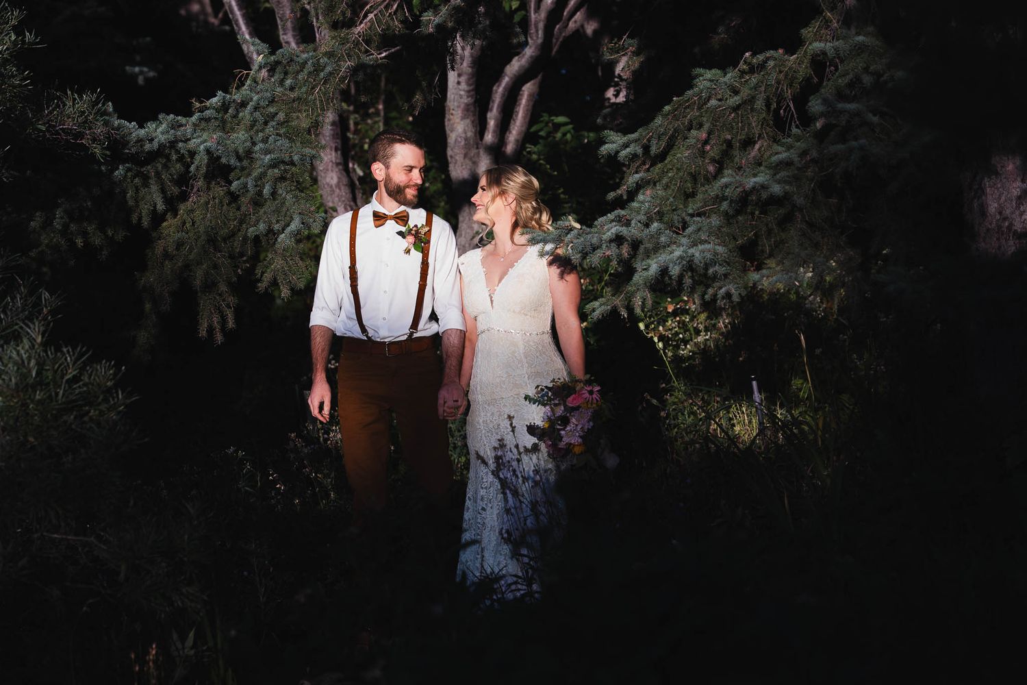 A couple in wedding attire stands together in a dark wooded area illuminated by dramatic lighting.