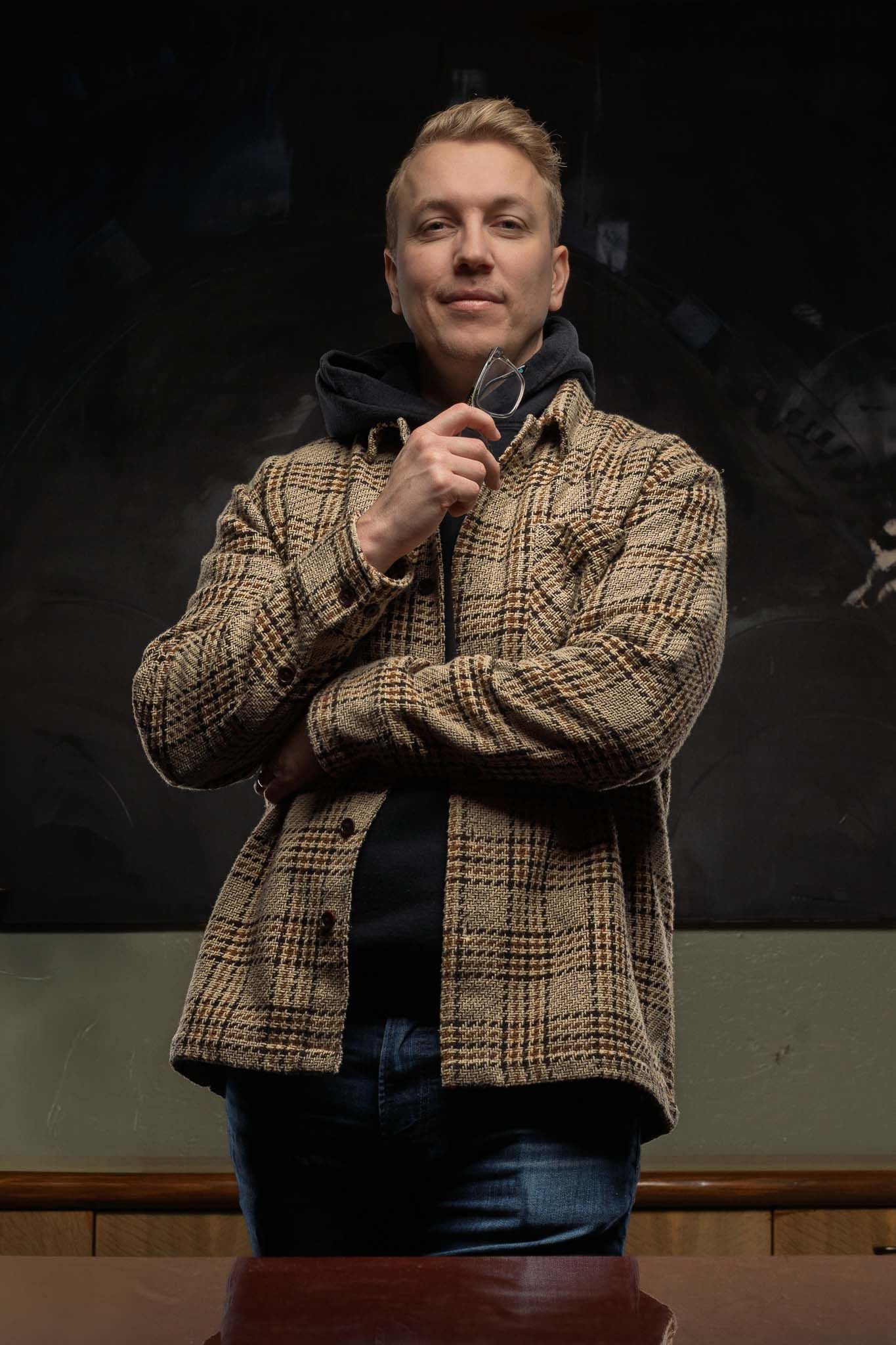 Person in brown plaid jacket poses against dark background with dramatic lighting.