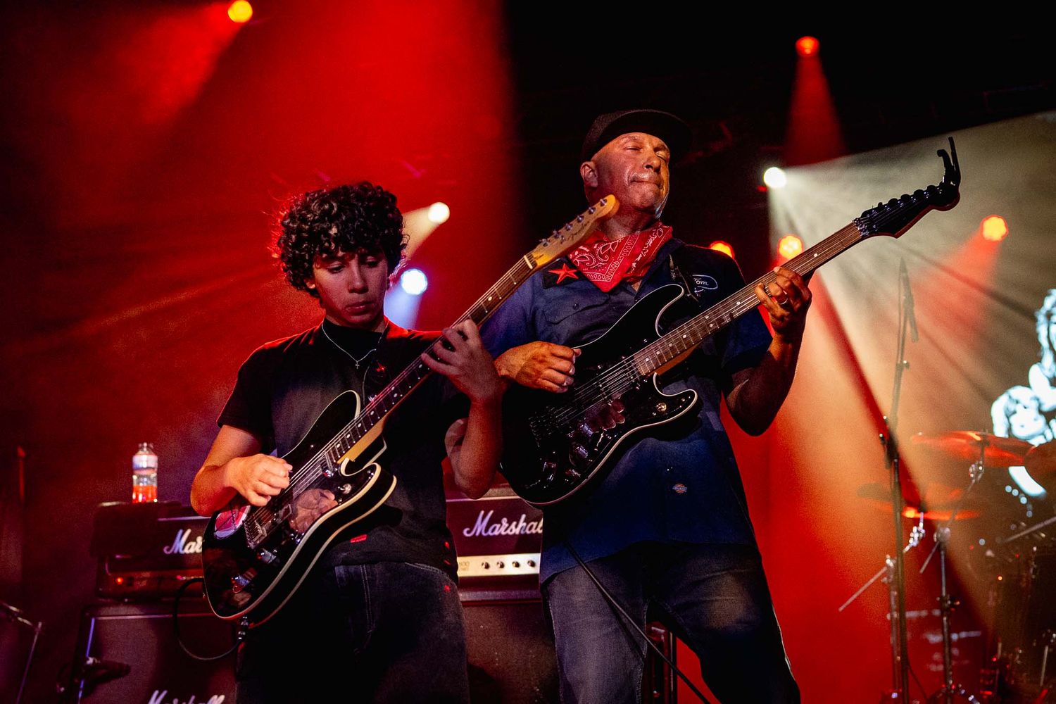 Tom Morello and Roman Morello performing on stage with red dramatic lighting and guitar players in silhouette.