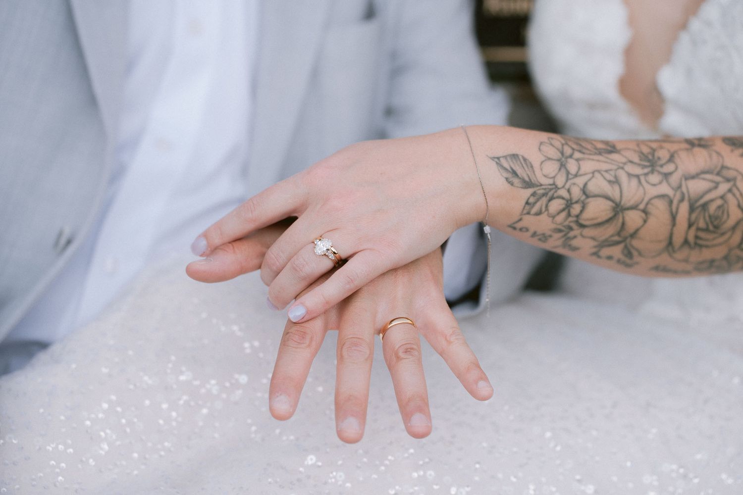 Close-up of hands with wedding rings and tattoos on white fabric.