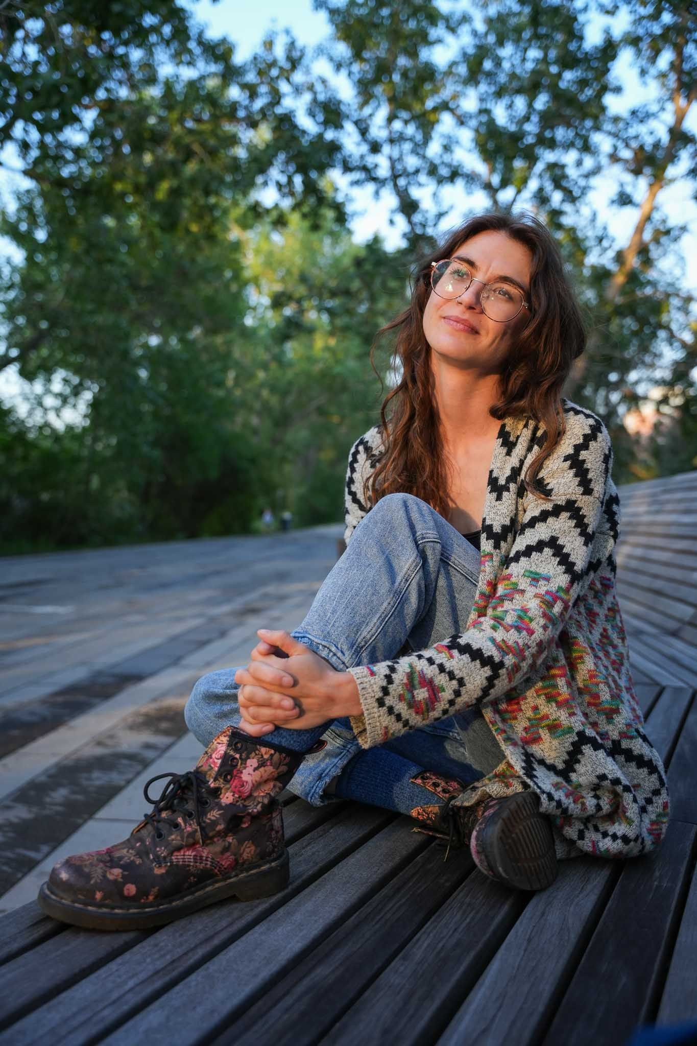 Person in patterned cardigan and jeans sitting on wooden deck surrounded by trees at sunset.