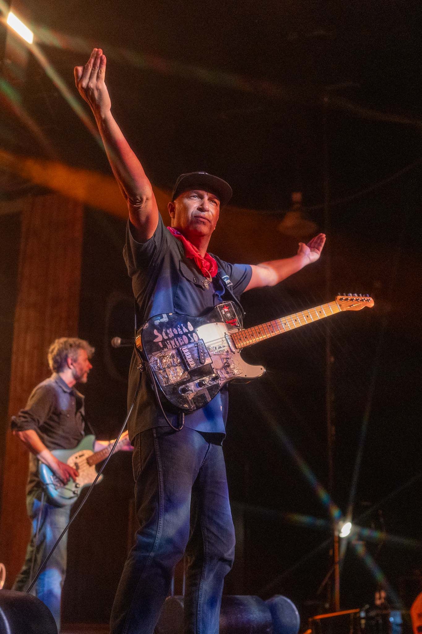 Tom Morello raise their arms while performing with guitars on stage under warm theatrical lighting.