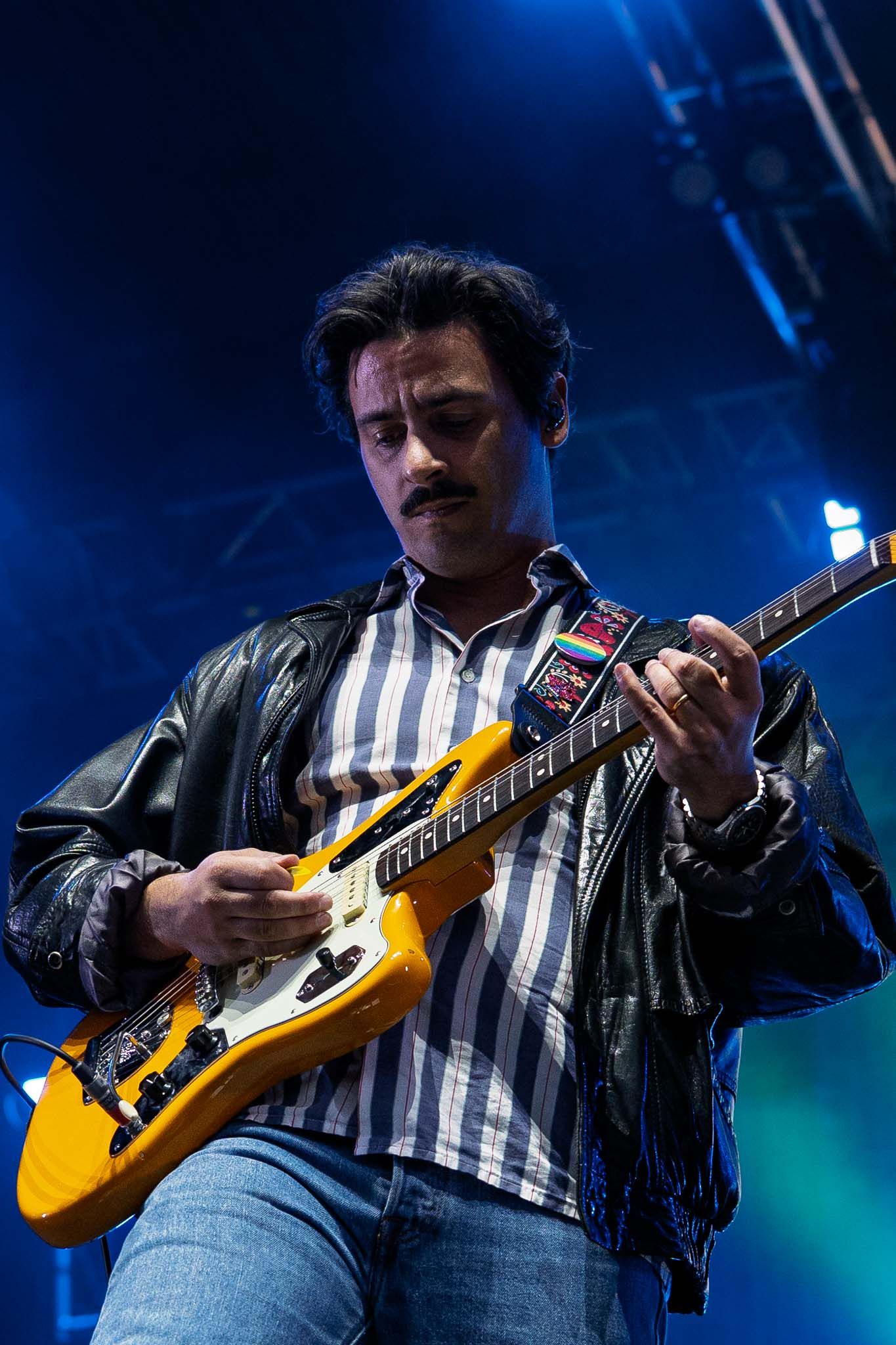 Guitarist in striped shirt playing yellow guitar under blue stage lights.