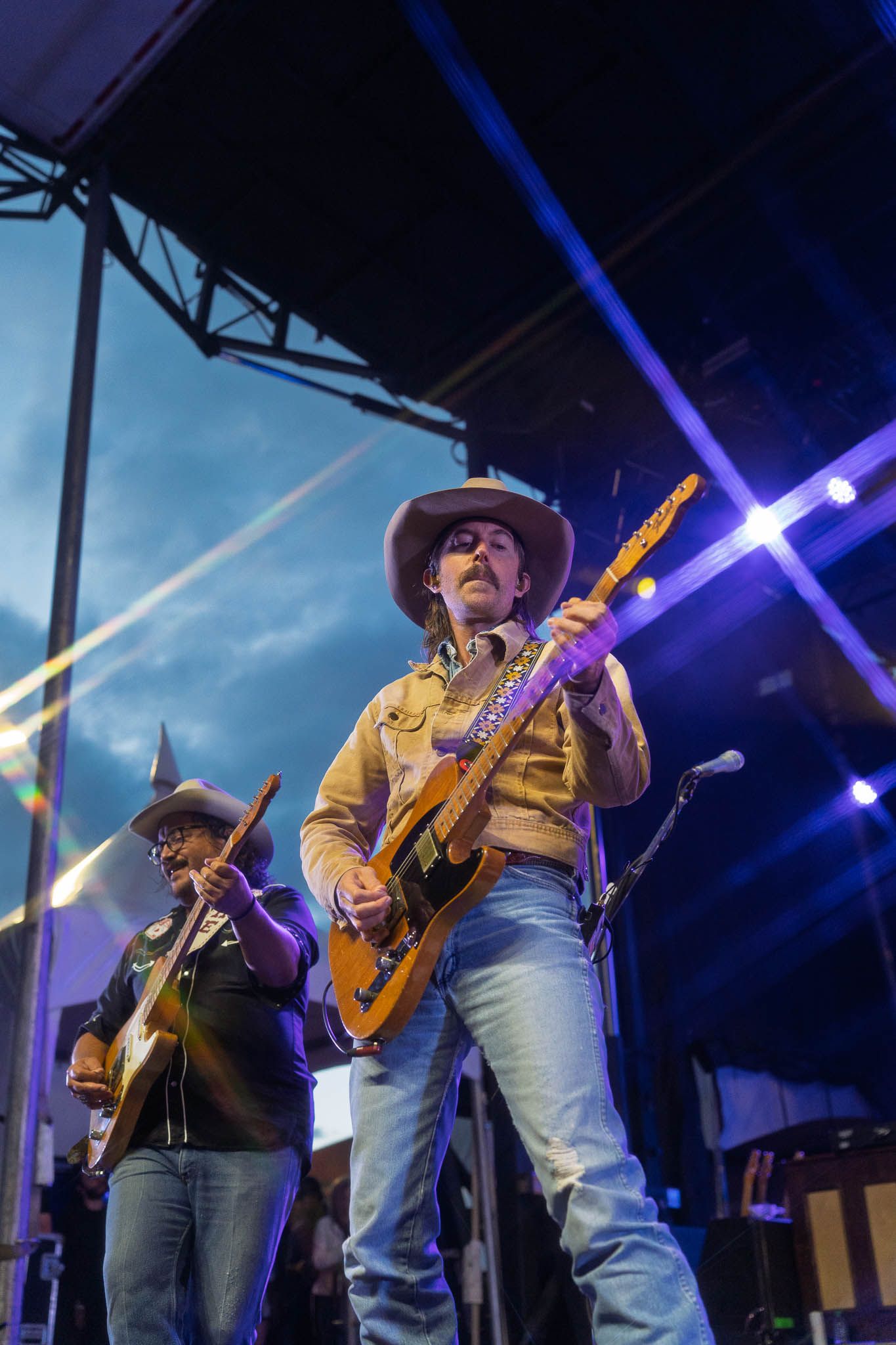 Musicians in western attire perform with guitars on outdoor stage with blue lighting effects.