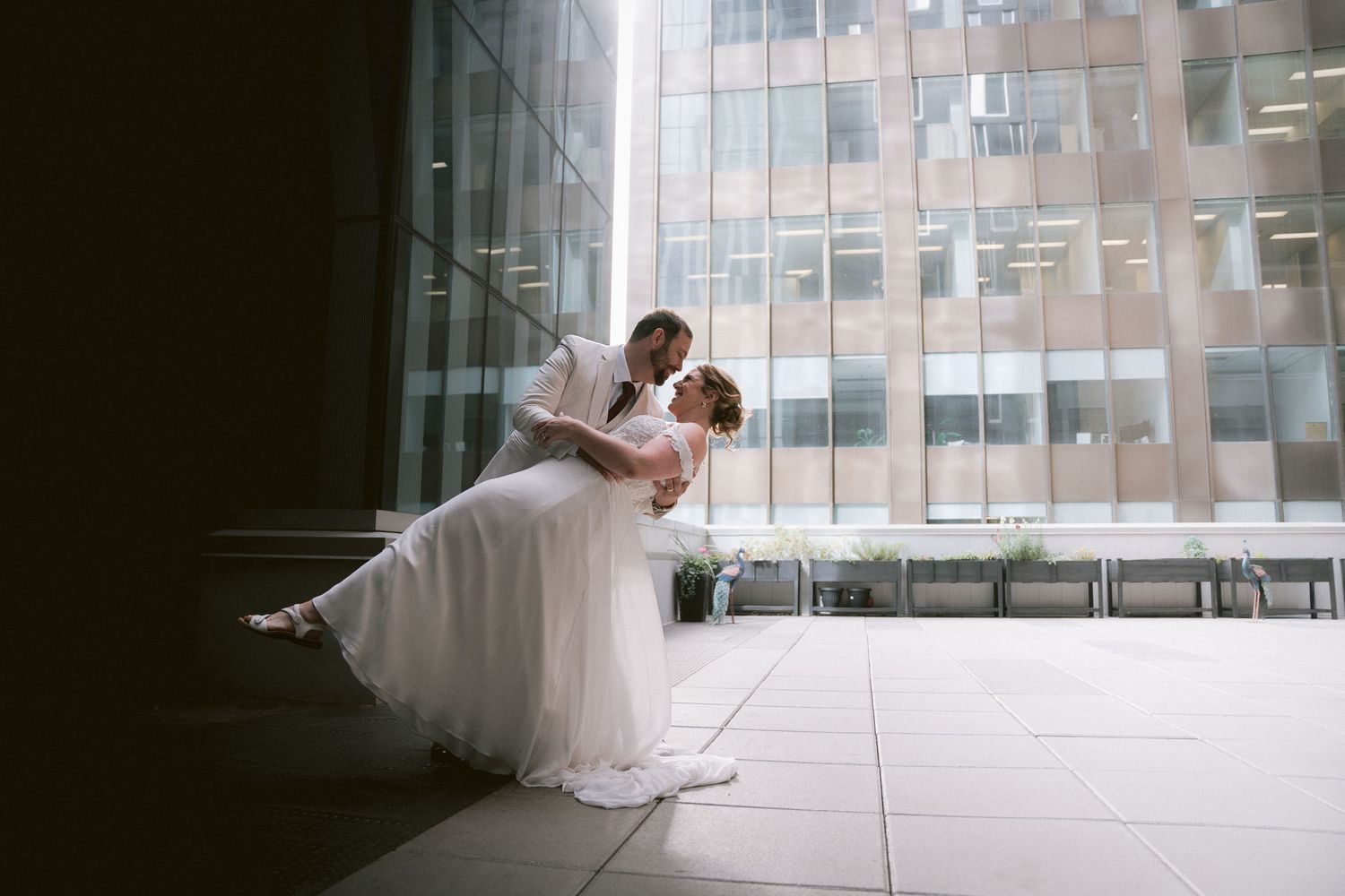 Wedding couple dances in modern architectural space with glass windows and urban backdrop.
