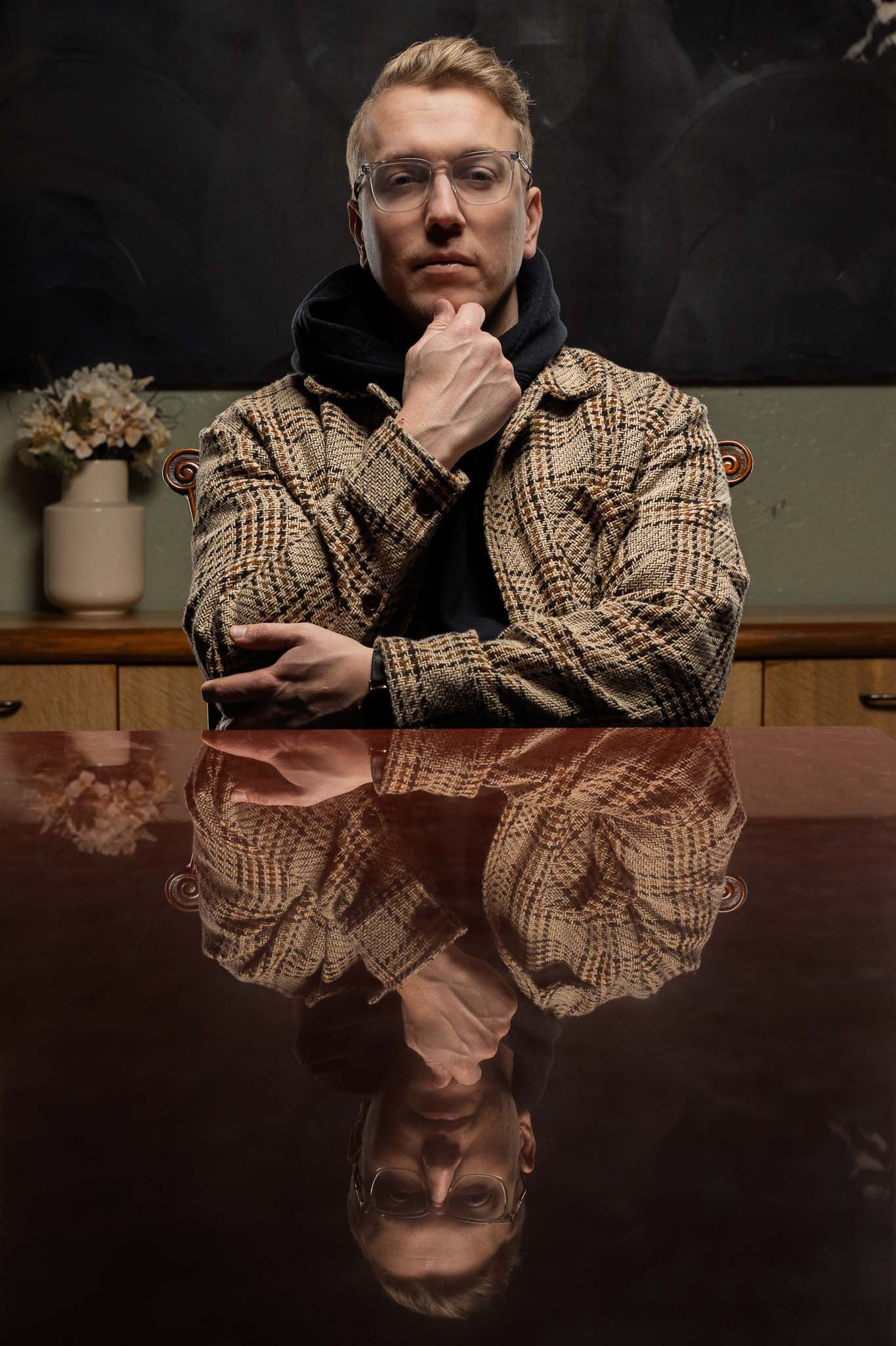 Person in plaid jacket poses artistically against reflective surface in moody lighting.