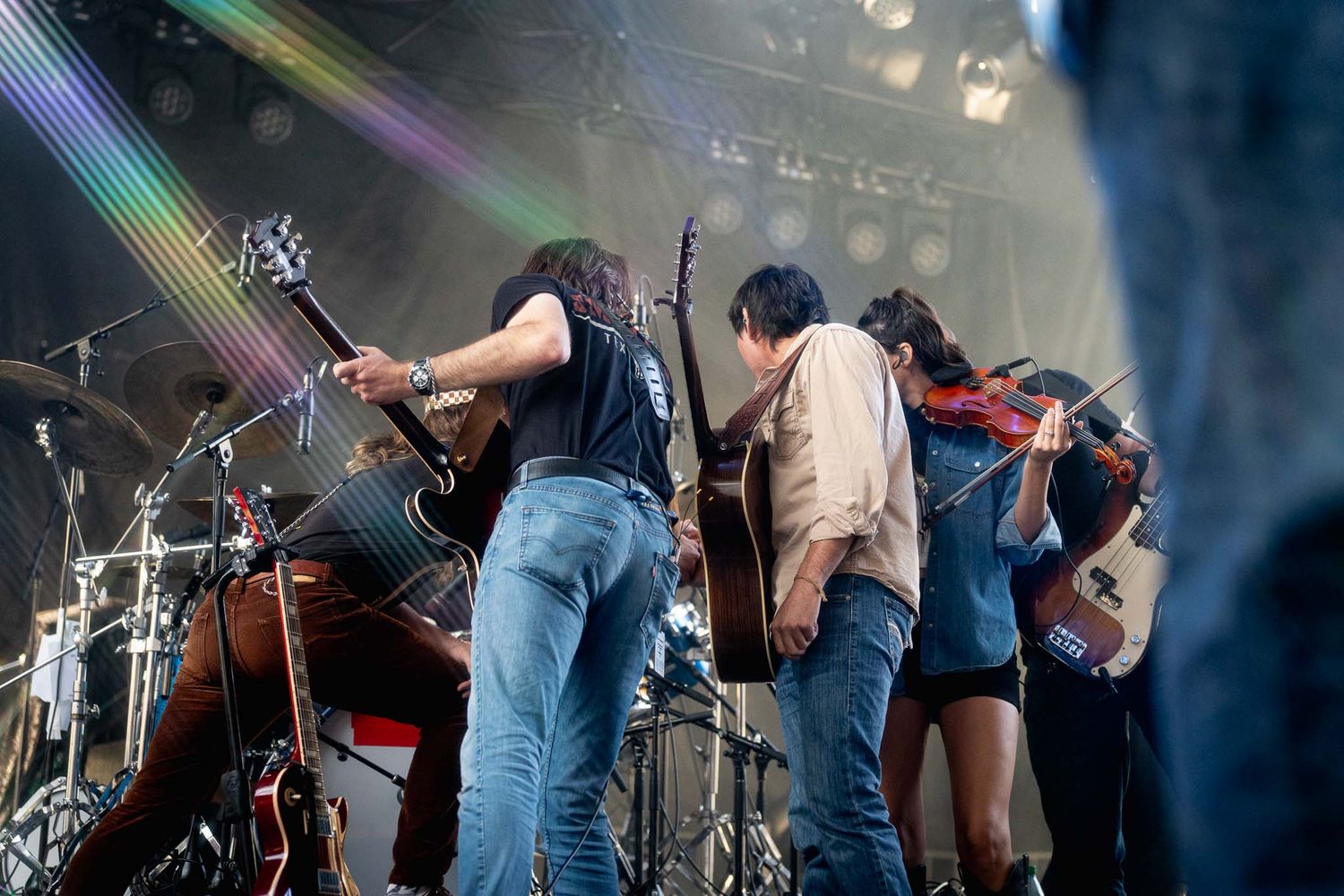 Musicians in jeans perform on stage with instruments under blue atmospheric lighting.