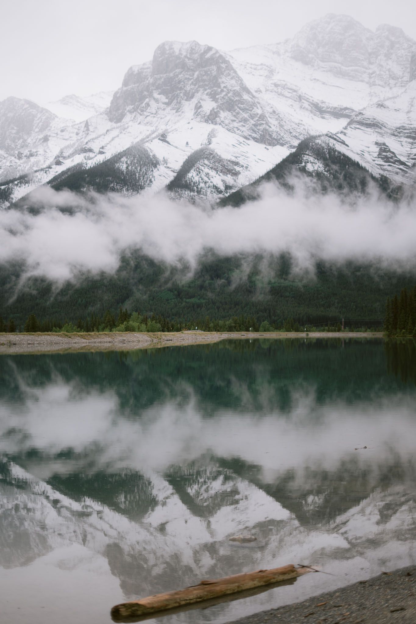 Misty mountain lake reflects snow-capped peaks with a fallen log in the foreground in Canmore, Alberta