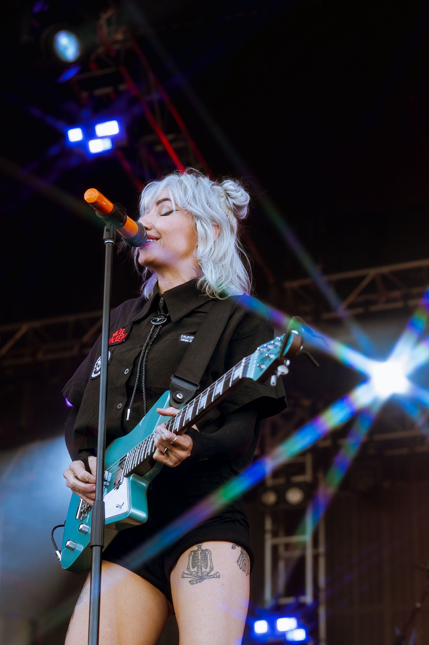 Singer Lights in black jacket sings into microphone while playing teal guitar on stage with dramatic lighting.