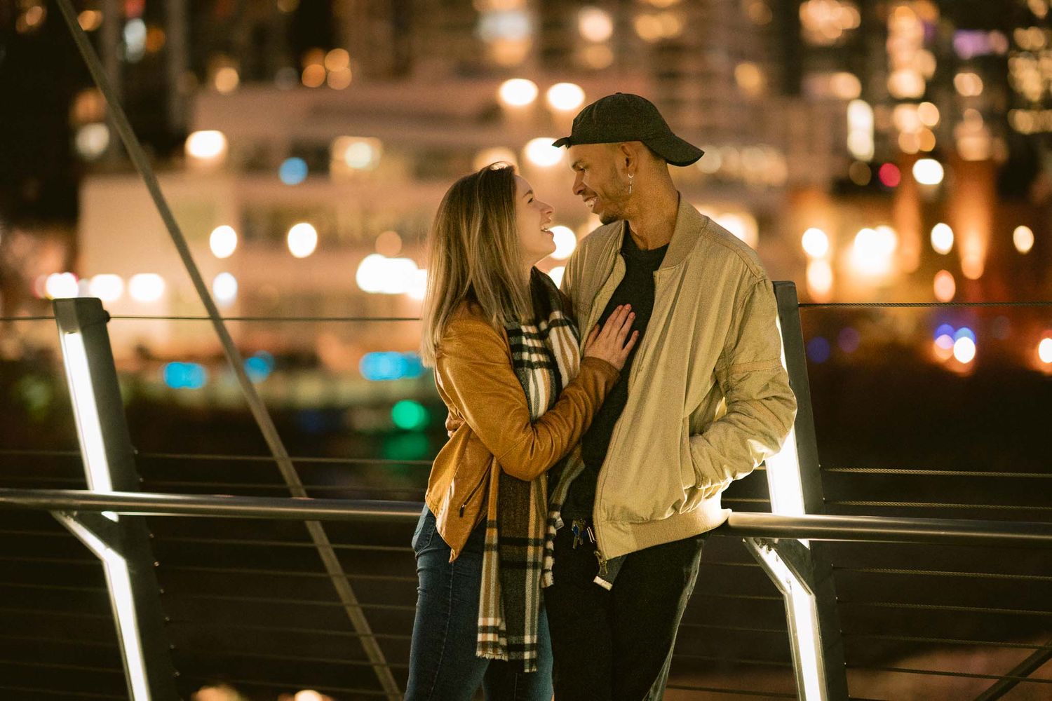A couple shares a romantic moment at night on a bridge with glowing lights in the background.