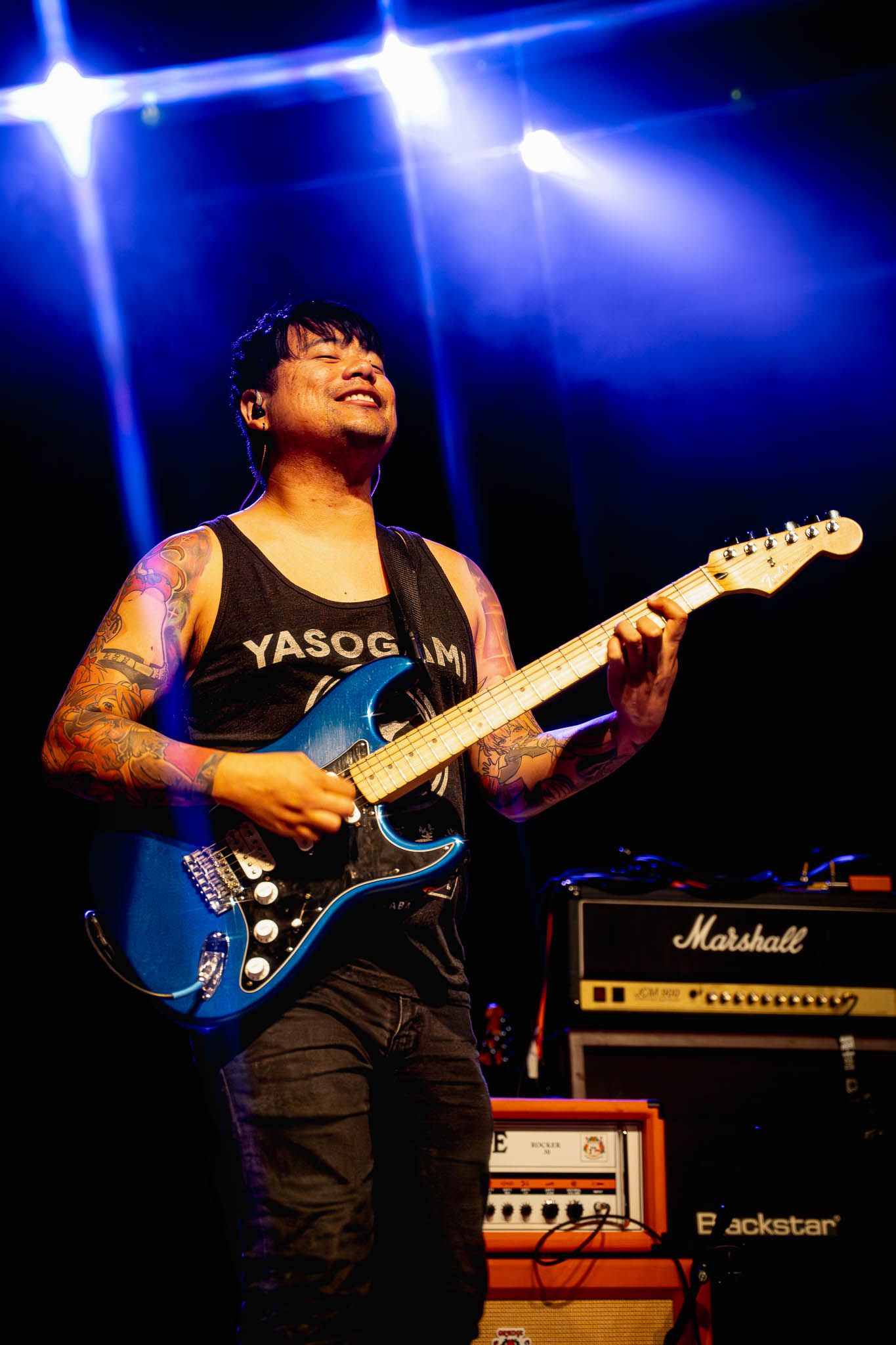Musician plays blue electric guitar against dark background with blue stage lighting and Marshall amp.