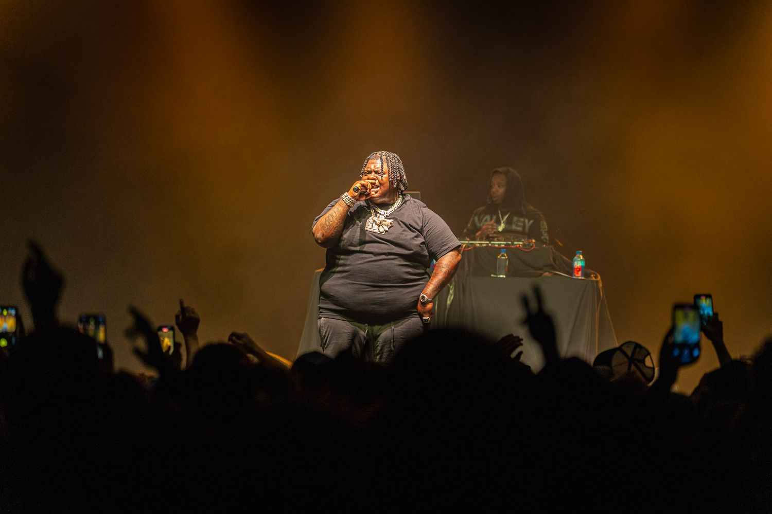 BigXthaPlug on stage with warm amber lighting and silhouetted crowd in foreground.
