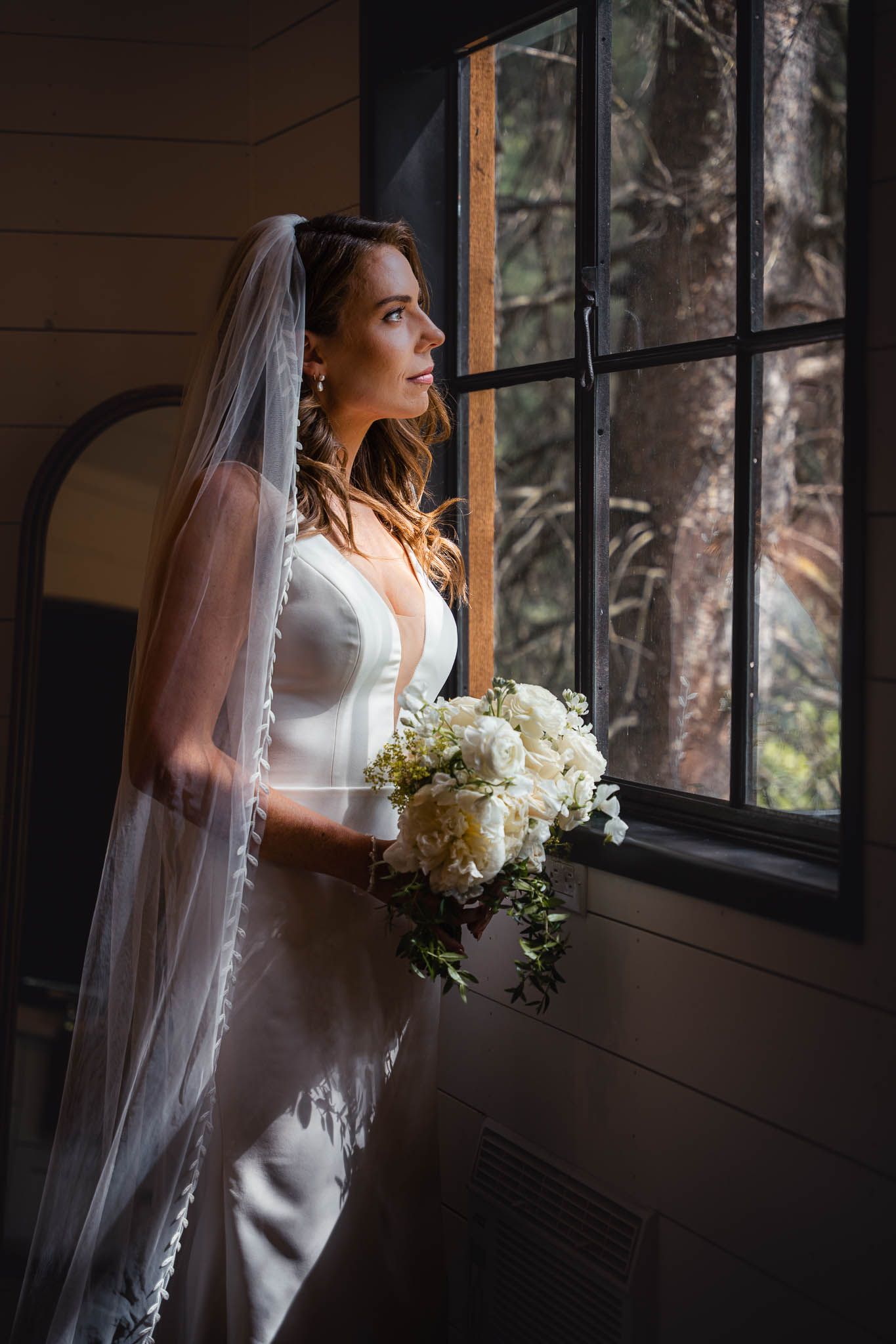 Bride in elegant white dress holds bouquet while standing by window in natural light in the Bridal Suite at The Valley Weddings