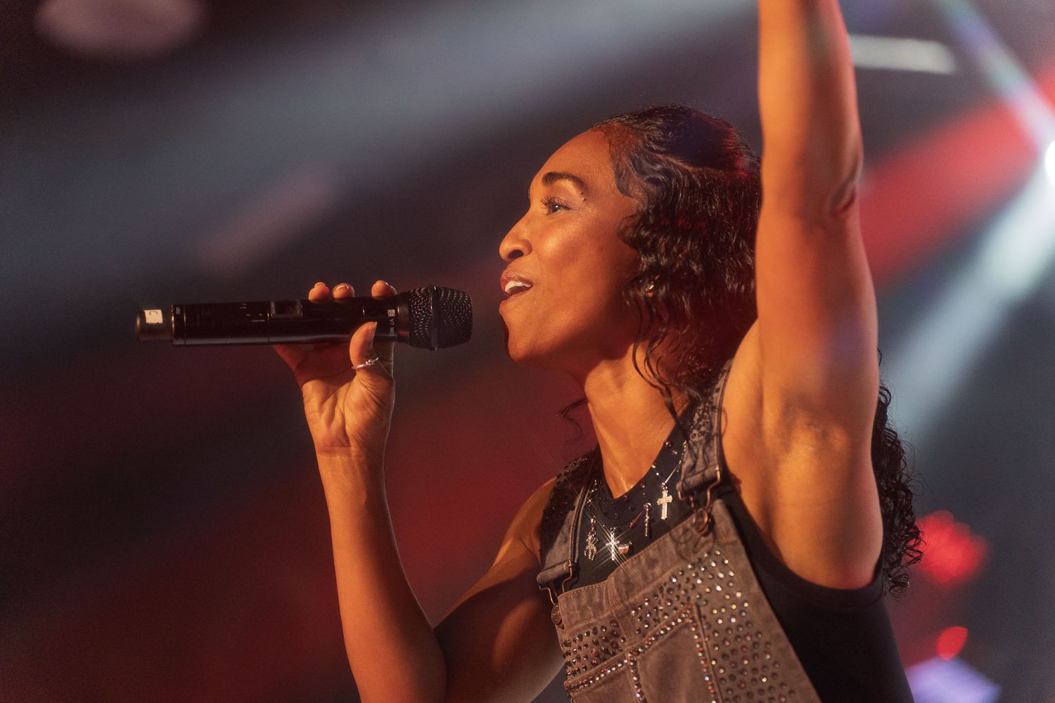 Performer in metallic outfit singing with raised arm under colorful stage lights.