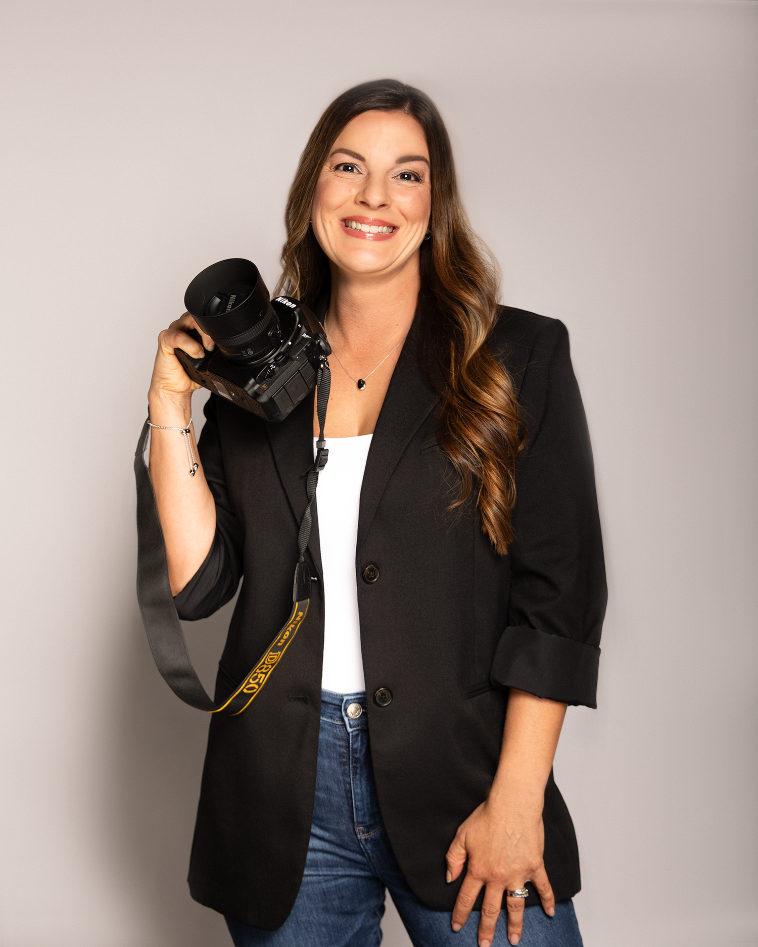 Professional female photographer smiling and holding a camera, wearing a black blazer and jeans against a gray backdrop.