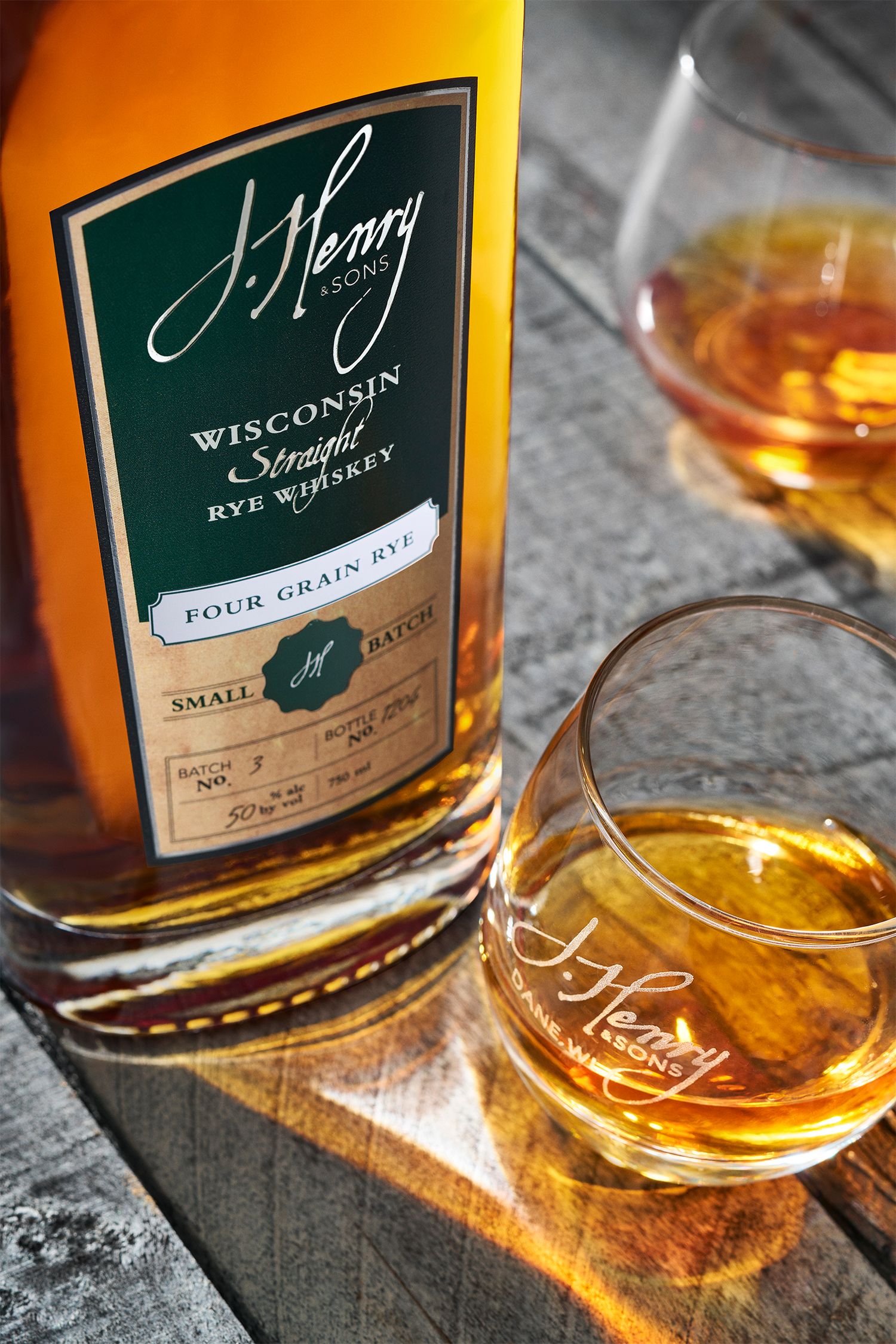 J. Henry & Sons Wisconsin Straight Rye Whiskey Four Grain bottle with two branded glasses on rustic wood surface.