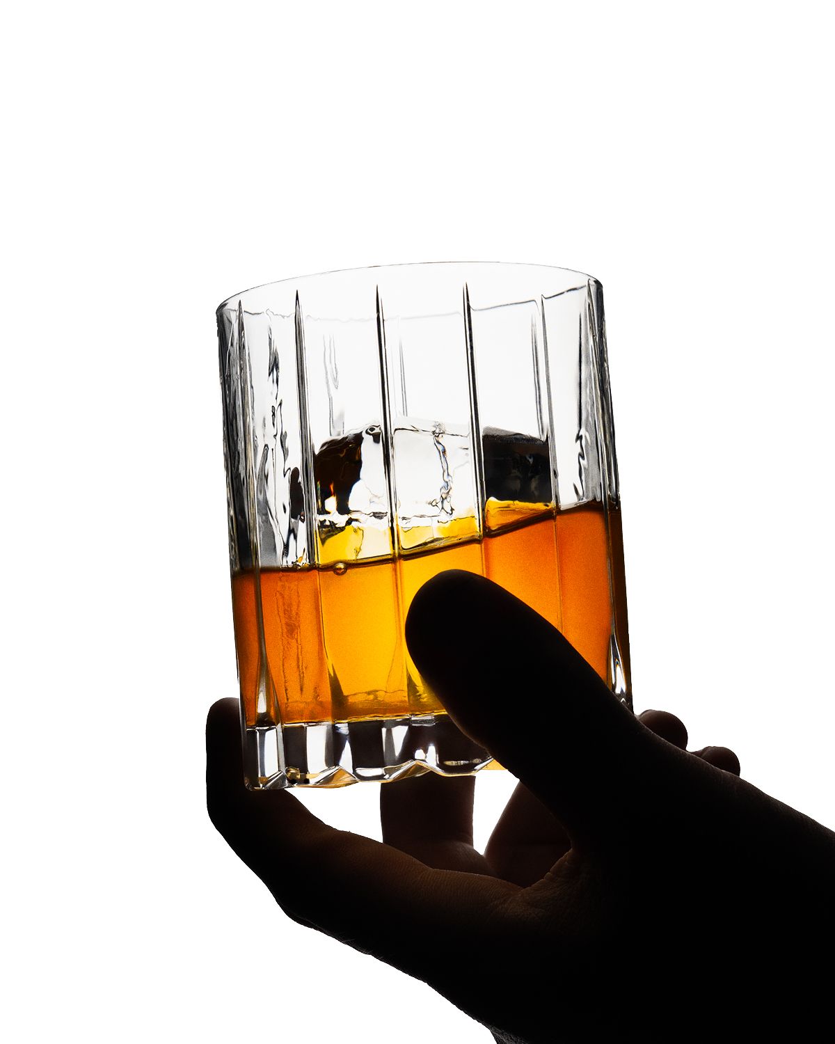 Silhouetted hand holding crystal glass of whiskey with ice against white background.