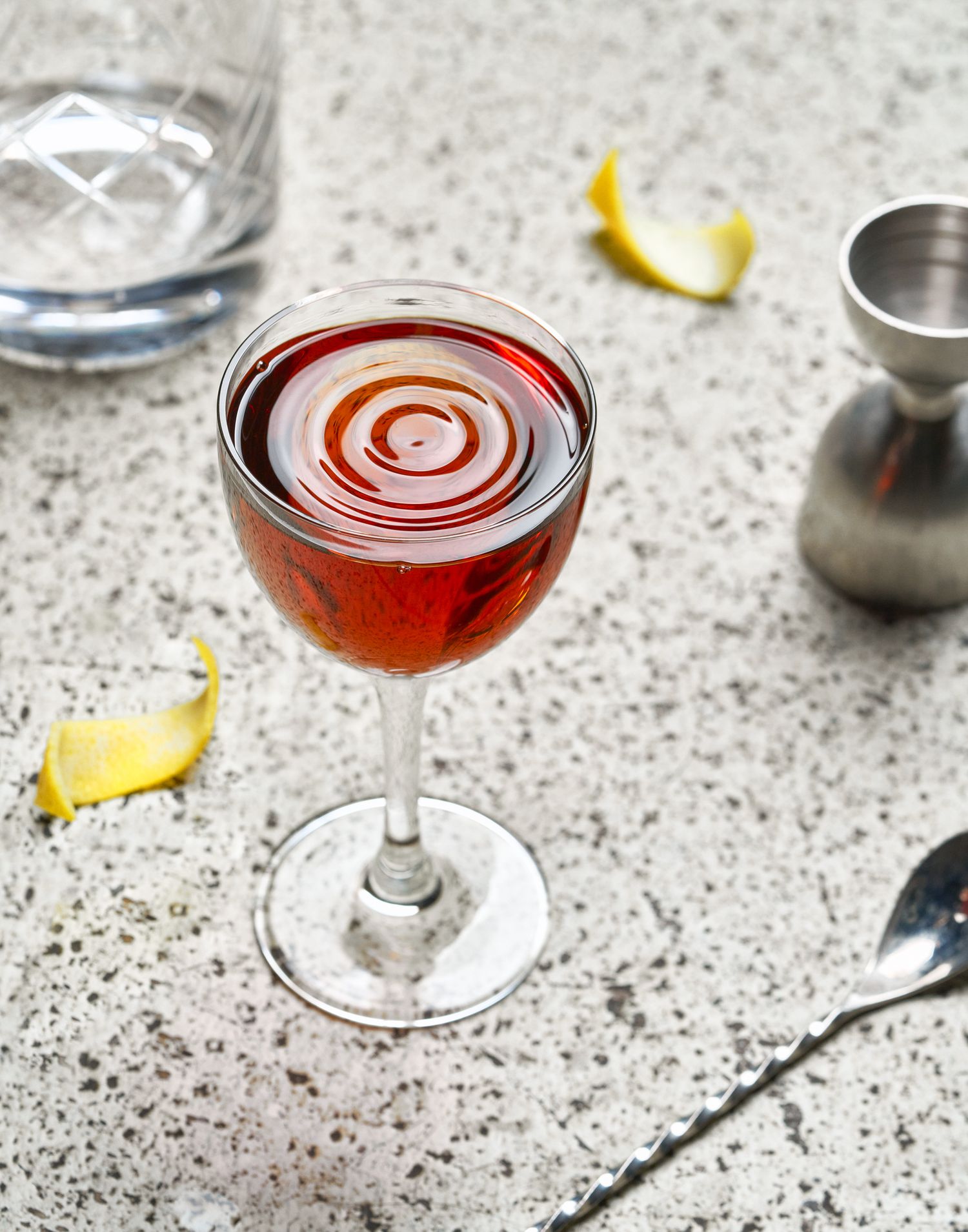 Red cocktail in martini glass with circular ripples on textured surface.