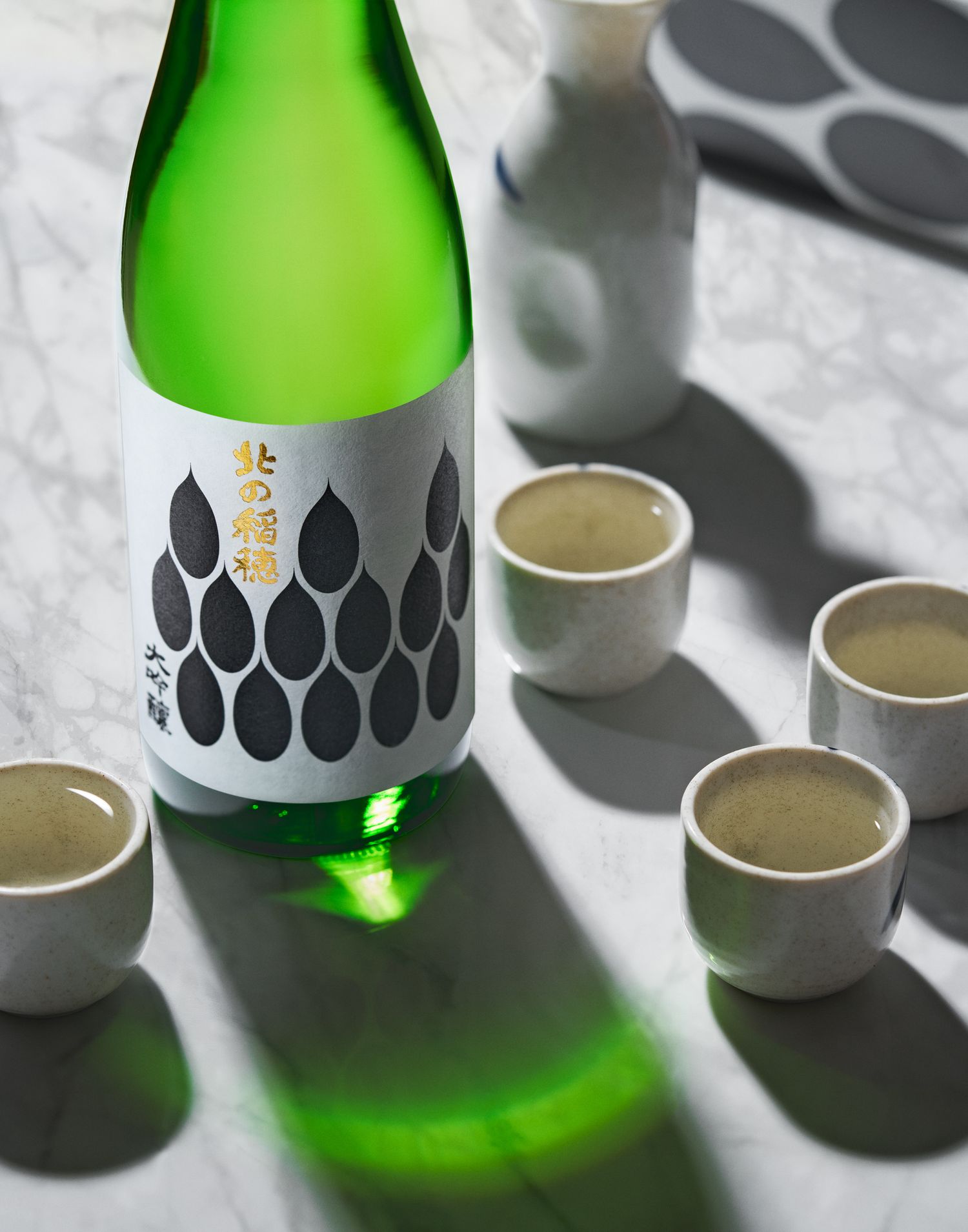 Green sake bottle with black geometric pattern design photographed with small ceramic cups.
