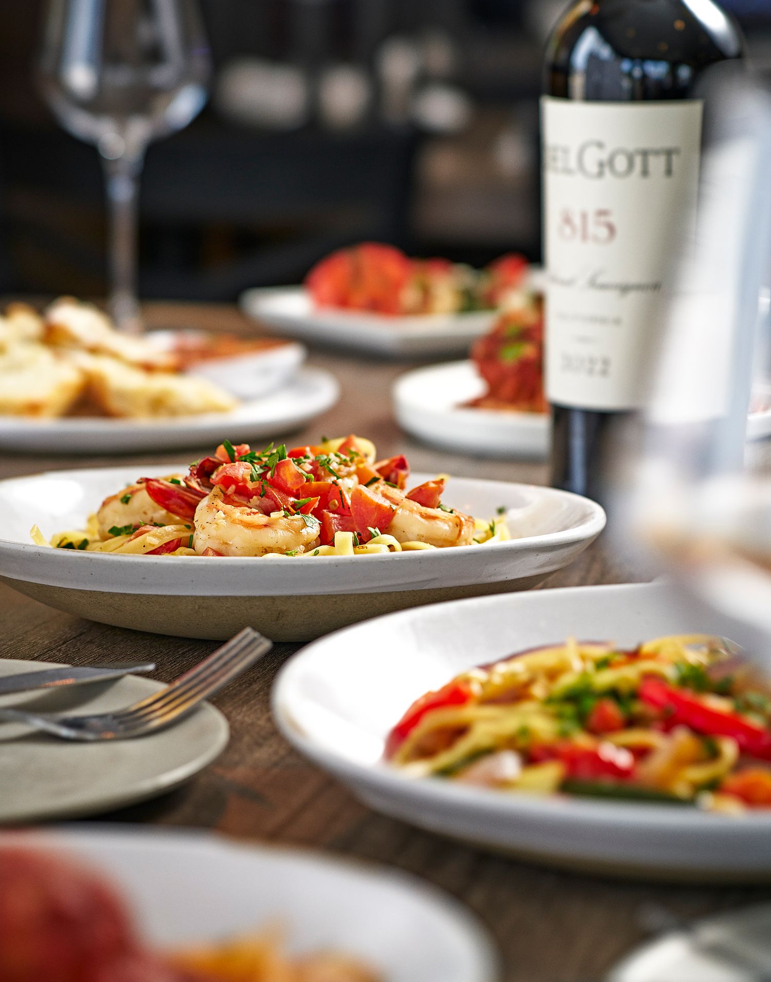 Close-up view of elegant plated pasta dishes with fresh ingredients and wine bottles on a dark wooden table.