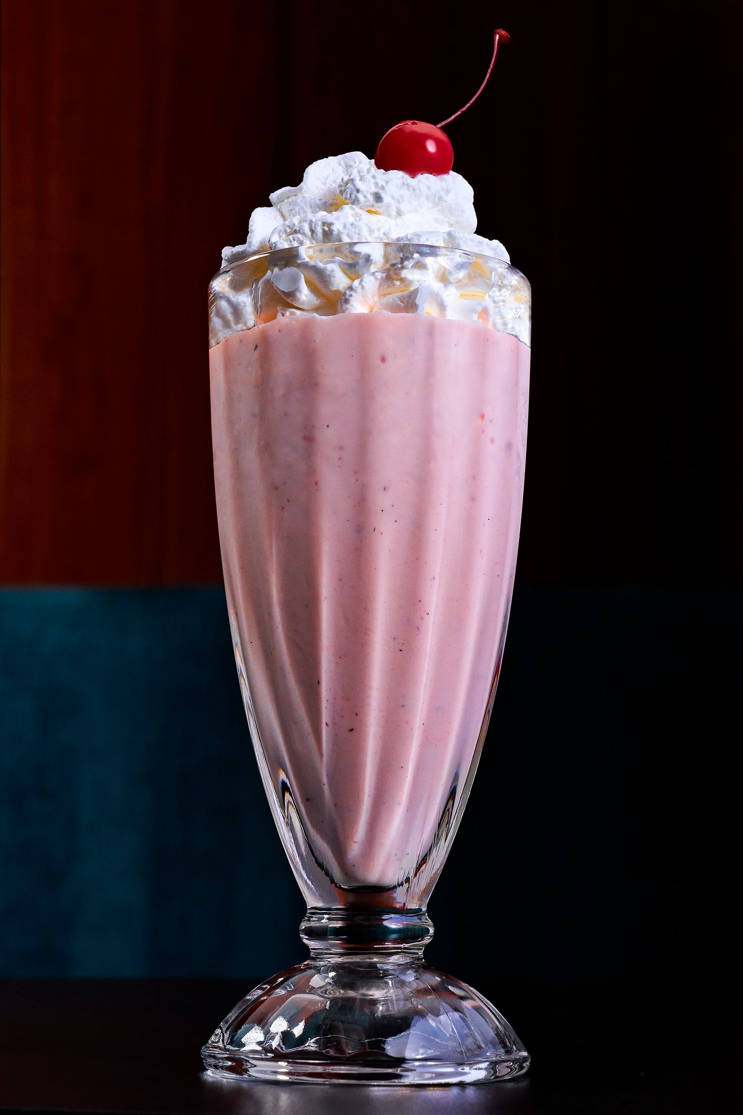 A pink strawberry milkshake topped with whipped cream and a cherry in a classic soda fountain glass.