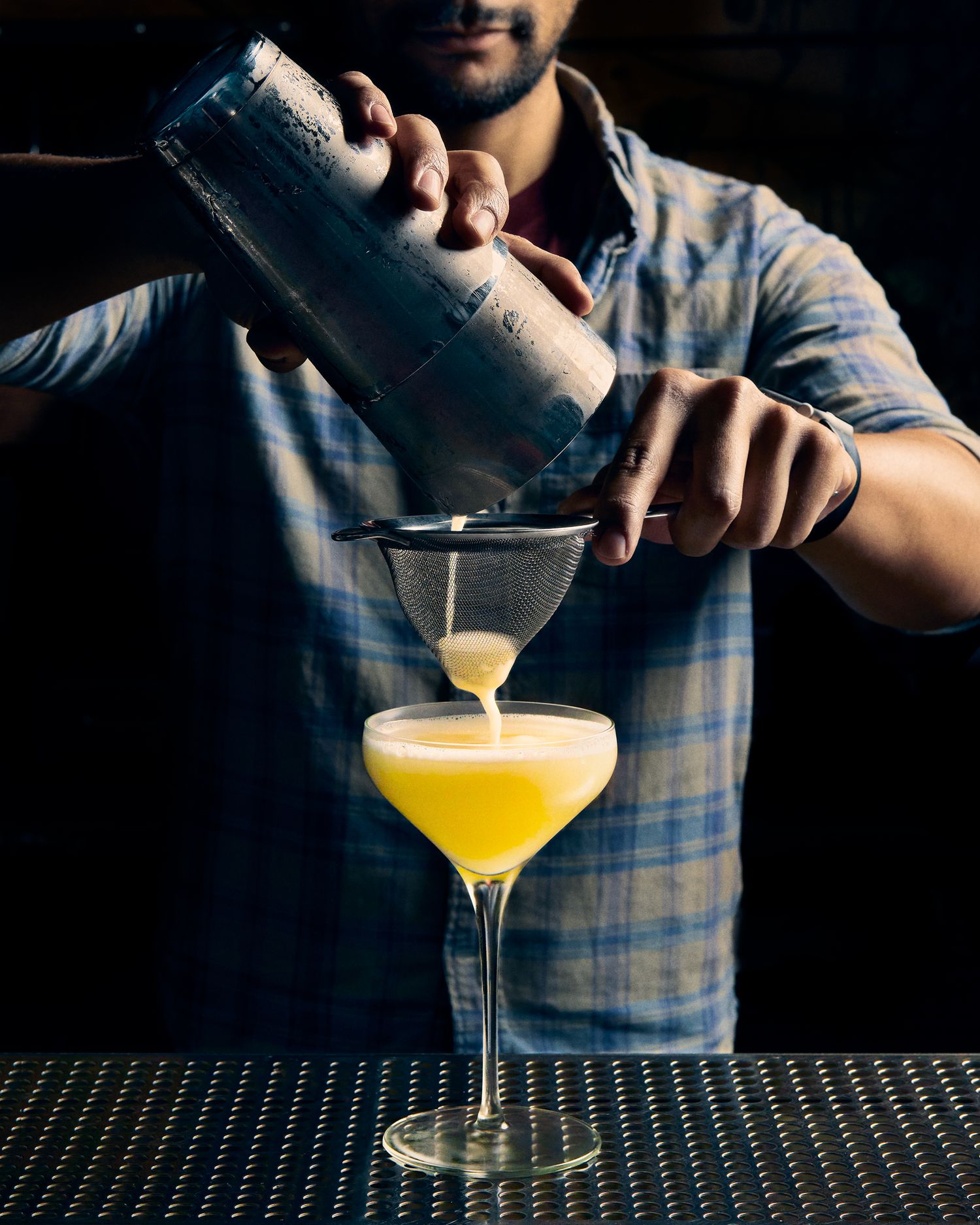 Bartender pouring a yellow cocktail into a coupe glass against a dark background.