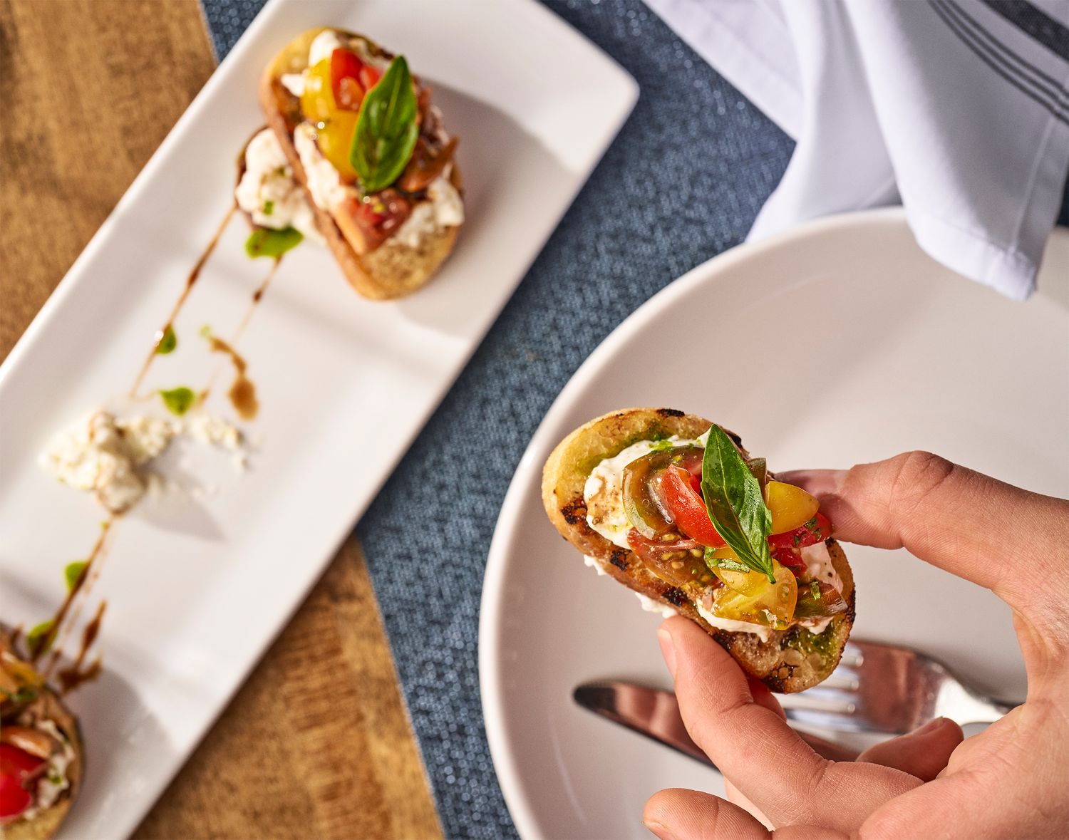 Hand holding bruschetta topped with fresh mozzarella, tomatoes, and basil on a white plate with serving platter behind.