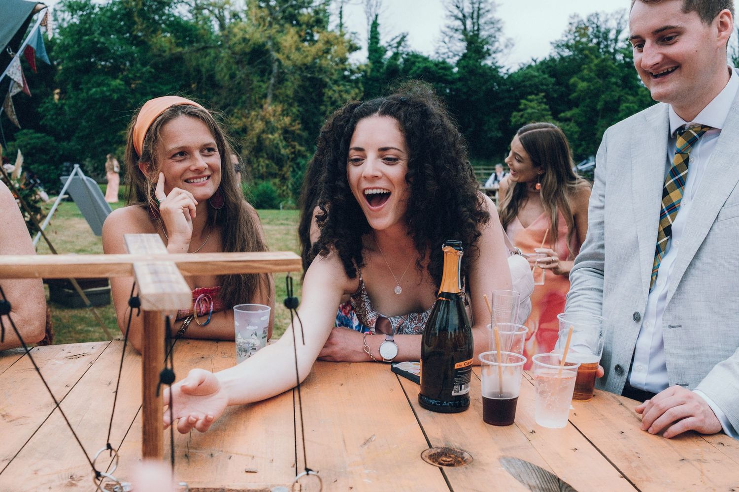 Wedding party gathers around rustic wooden tables for drinks and celebration.