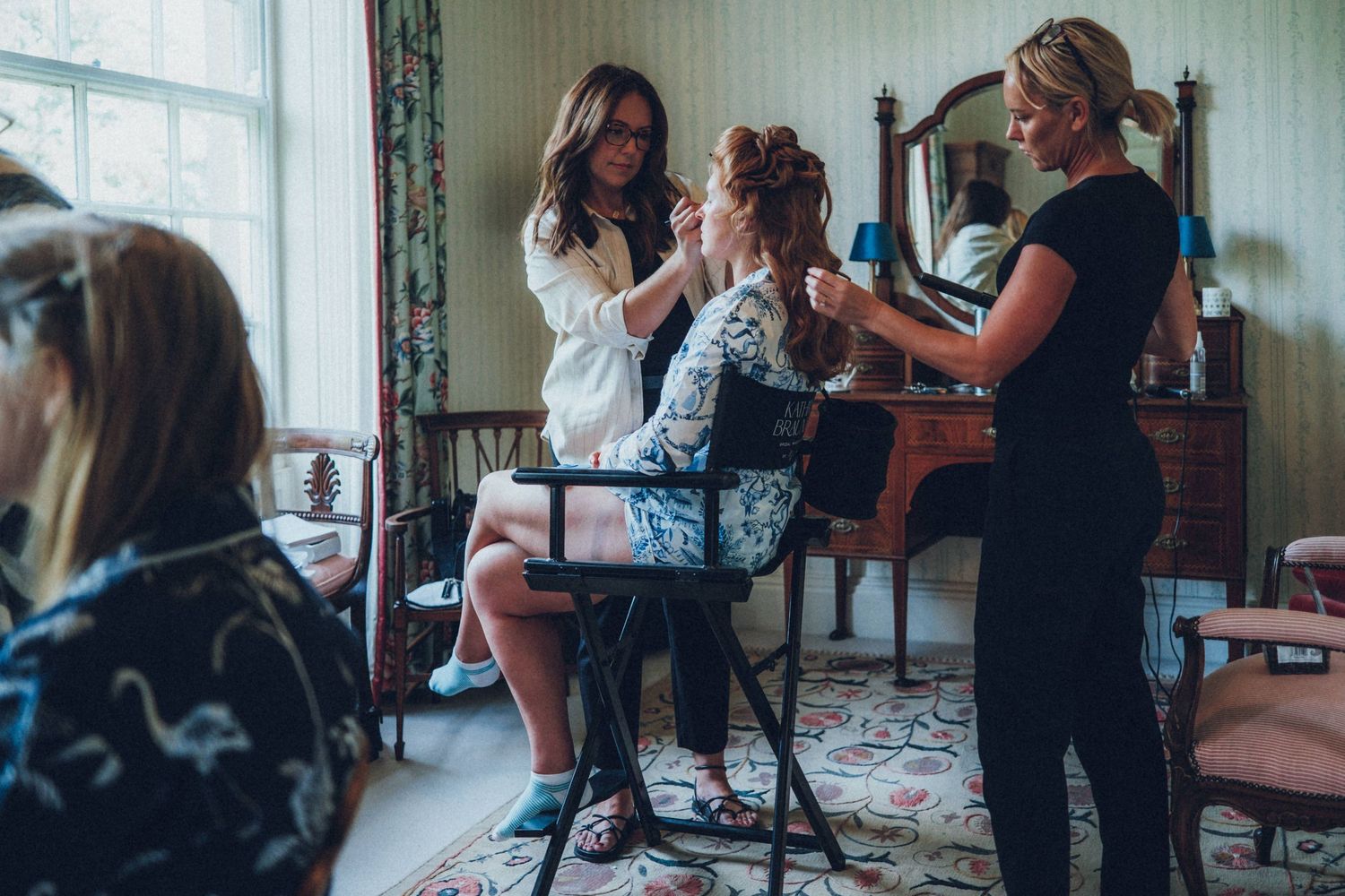 Hairstylists preparing a bride for her wedding day in a sunlit room with vintage decor.