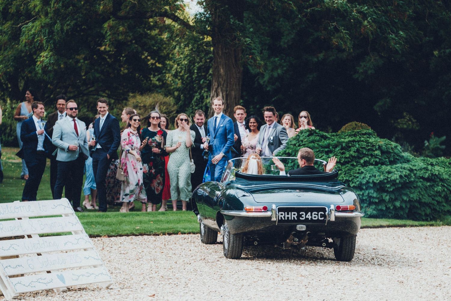 Wedding guests gather around vintage cars on gravel driveway.