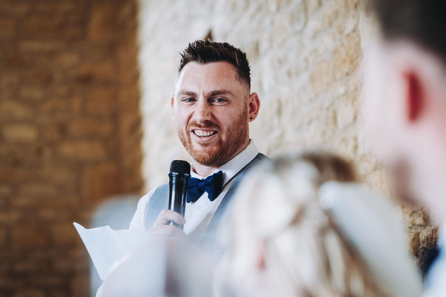 A person wearing a black bowtie gives a heartfelt wedding speech with a microphone against exposed brick walls.