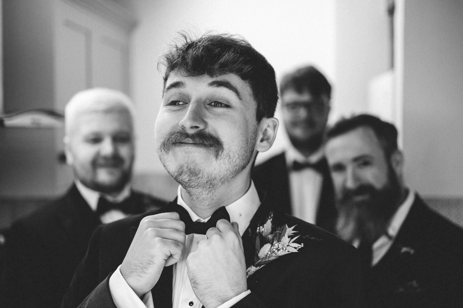Black and white photo of a groom adjusting his bowtie while groomsmen look on with smiles at a wedding.