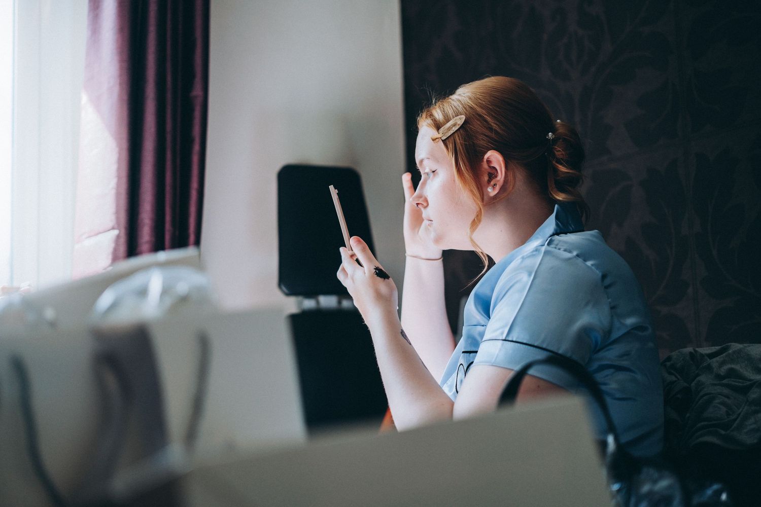 Someone sits on a couch in blue shirt looking at their phone while vaping or smoking in a dimly lit room.